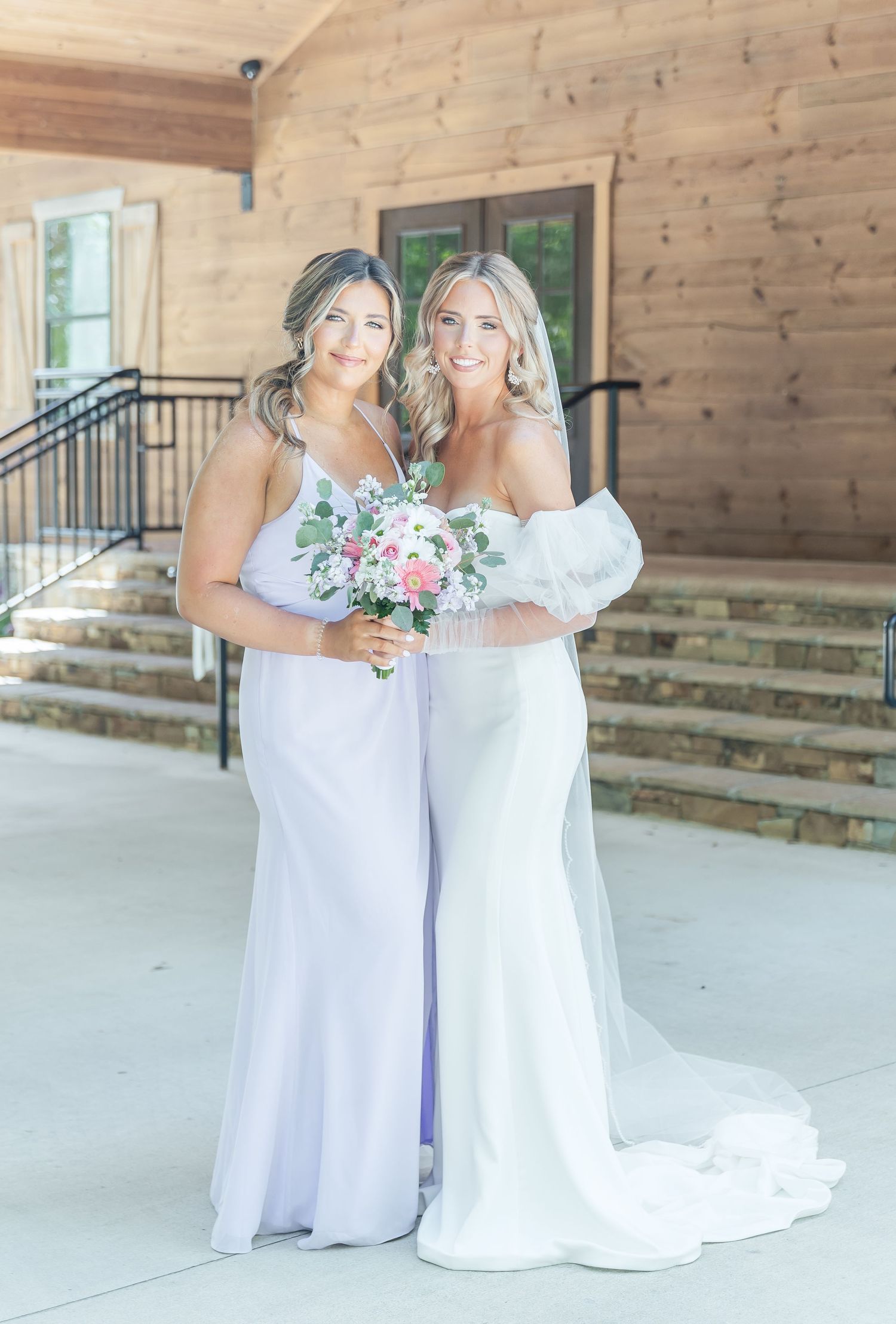 Bride and bridesmaid in white gowns pose together with matching pink and white bouquets.
