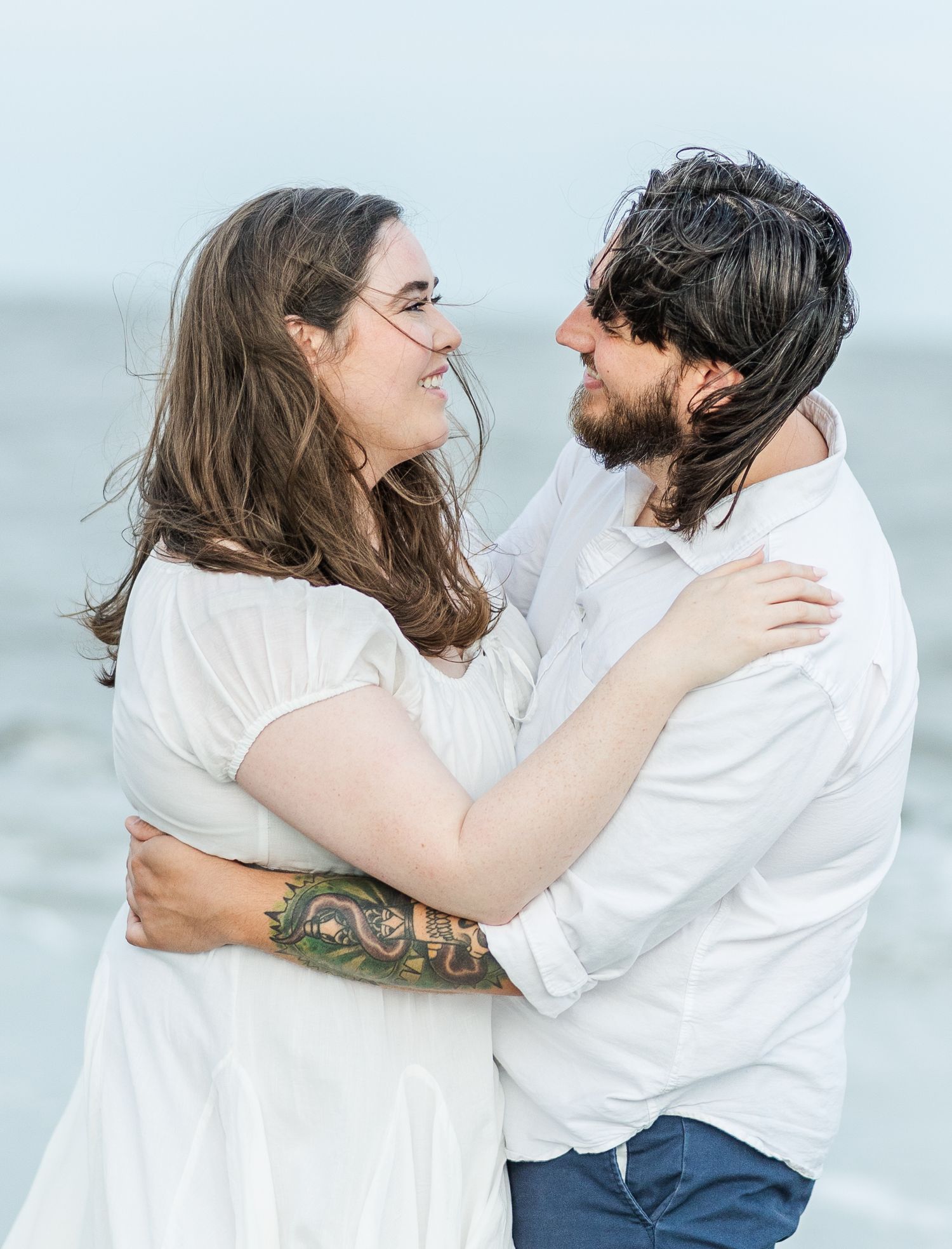 A close-up portrait of a couple in white clothing smiling at each other on the beach.