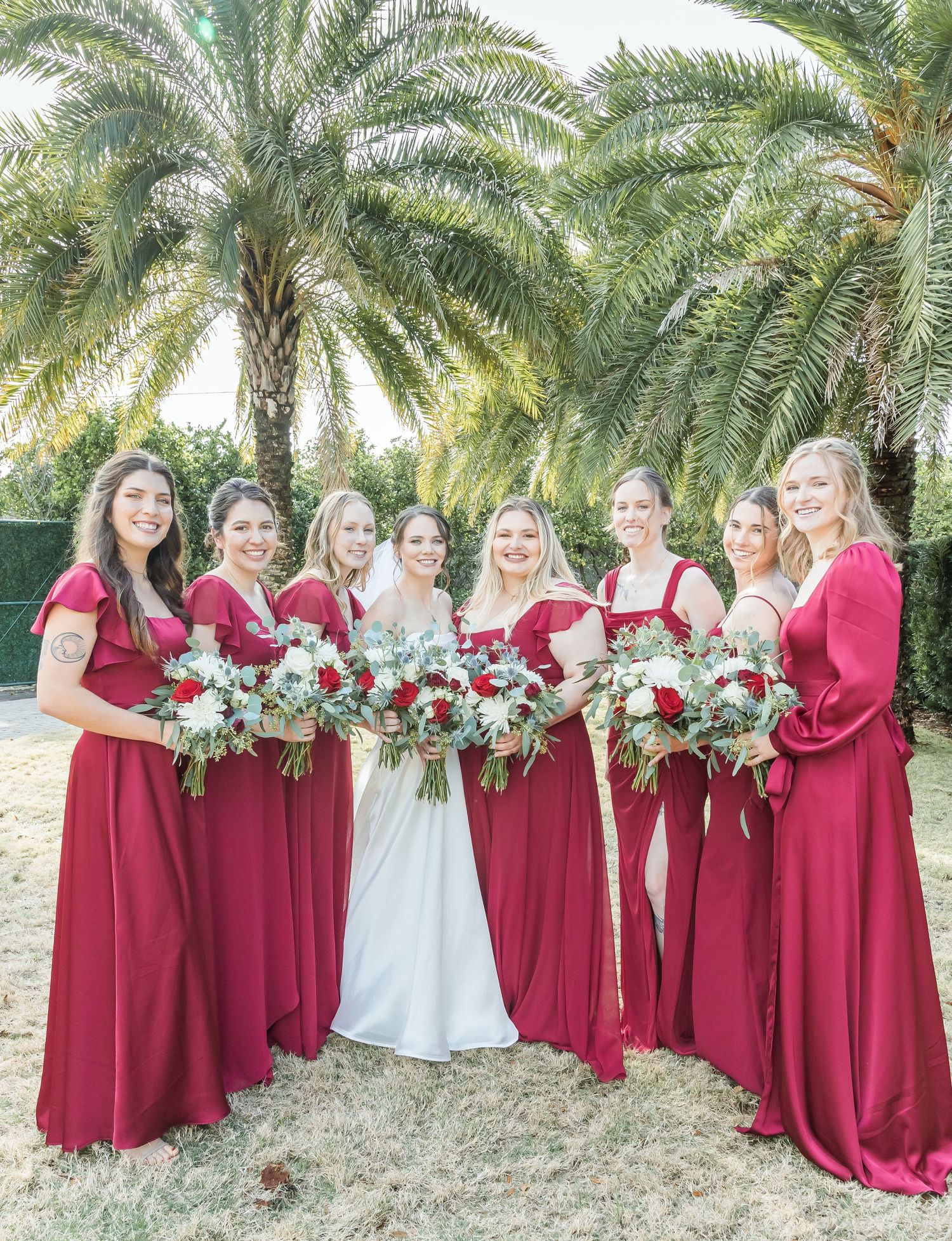 Bridesmaids in burgundy dresses pose with white bouquets against palm tree backdrop.