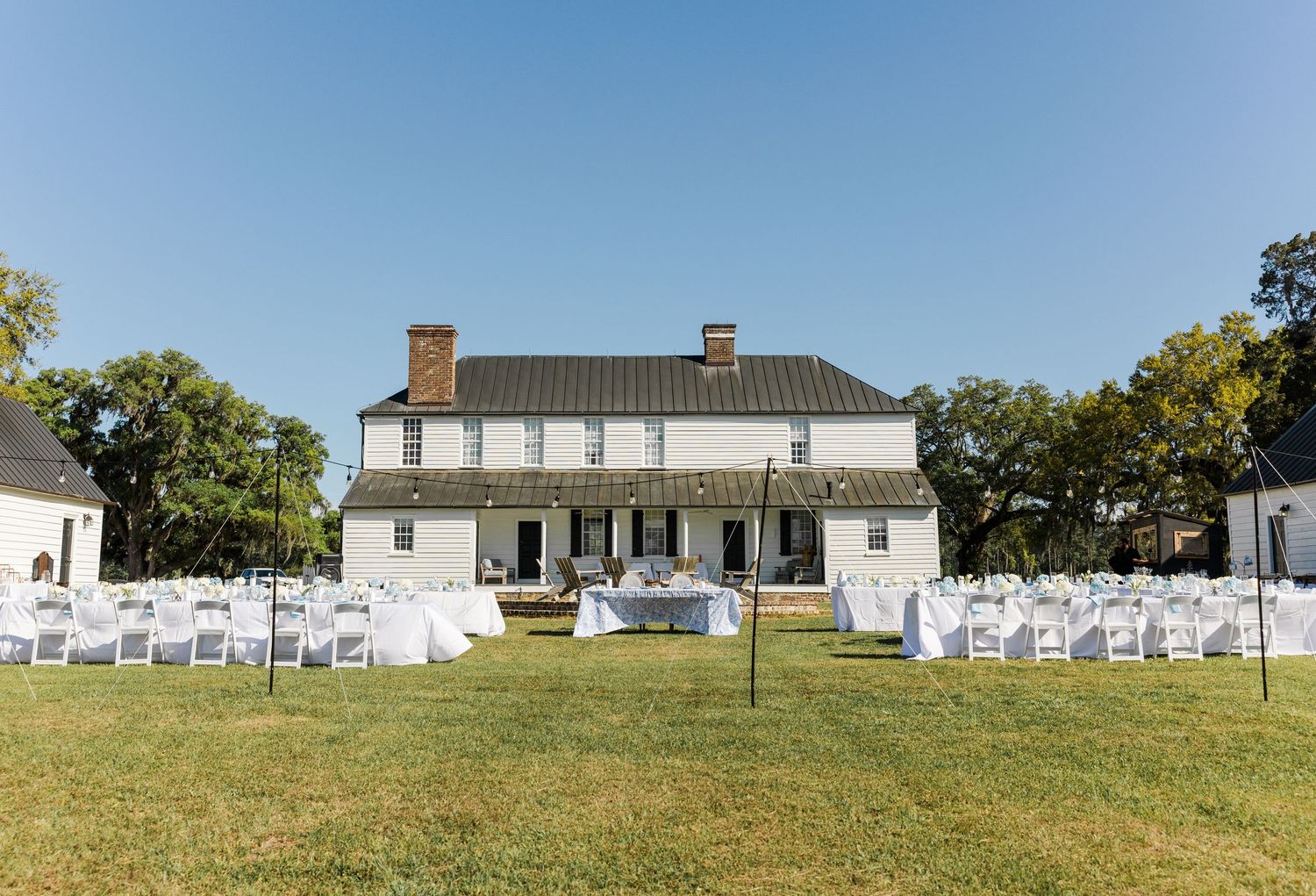 White colonial manor house with outdoor wedding ceremony chairs arranged on green lawn under blue sky.