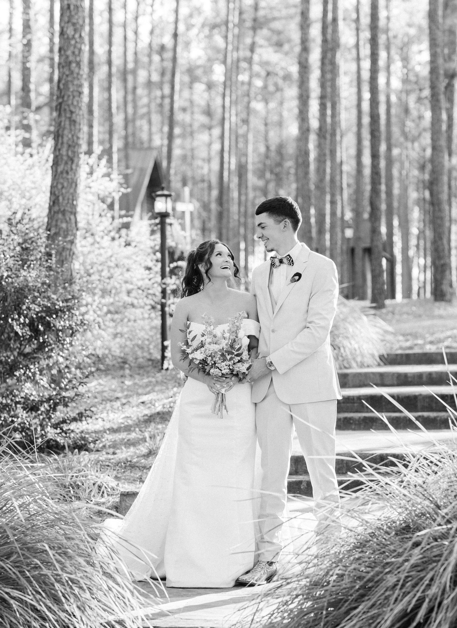 Black and white photo of smiling bride and groom walking together among tall pine trees at outdoor wedding venue.