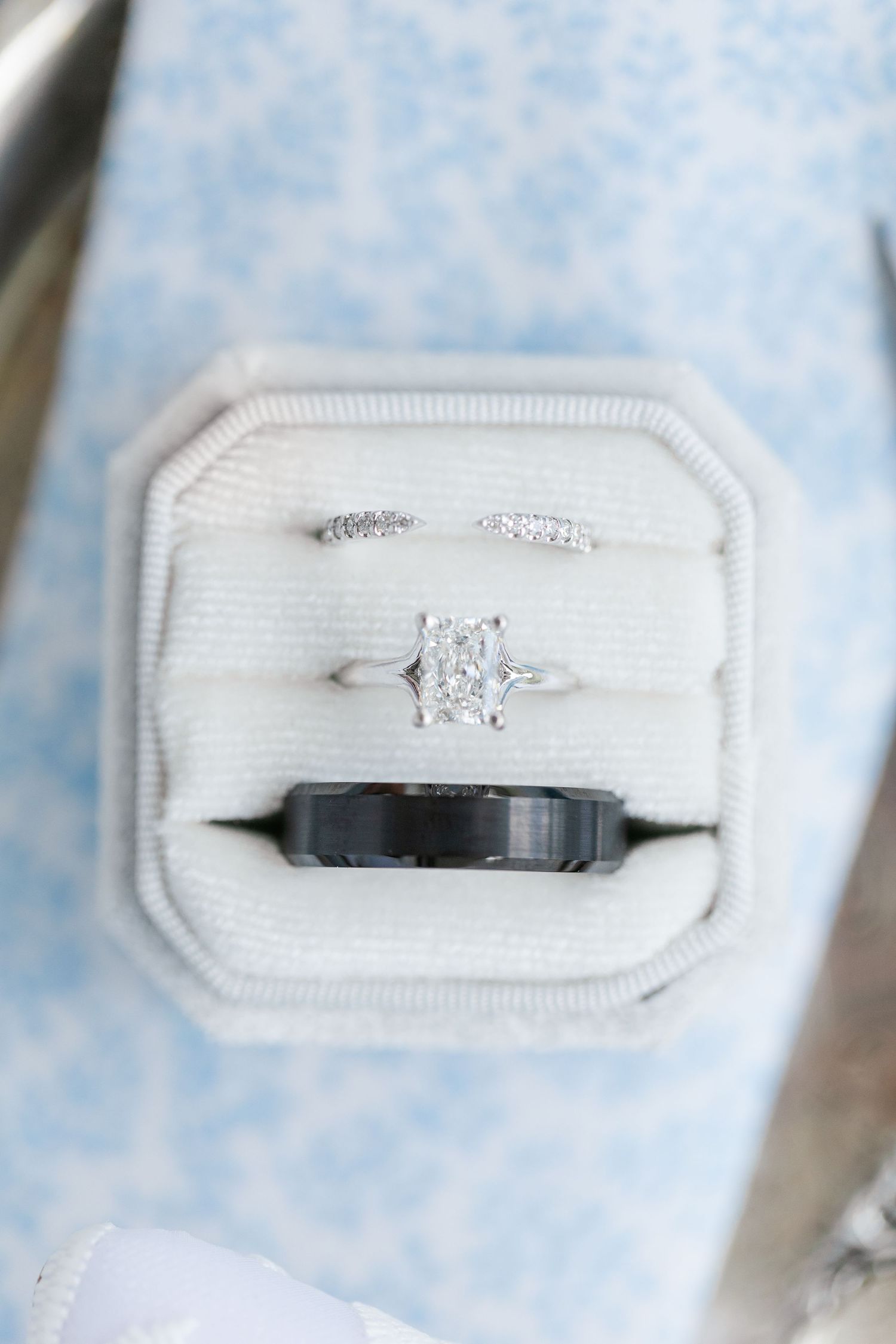 Wedding ring set in cream velvet box featuring radiant cut diamond engagement ring, diamond band, and black tungsten groom band.