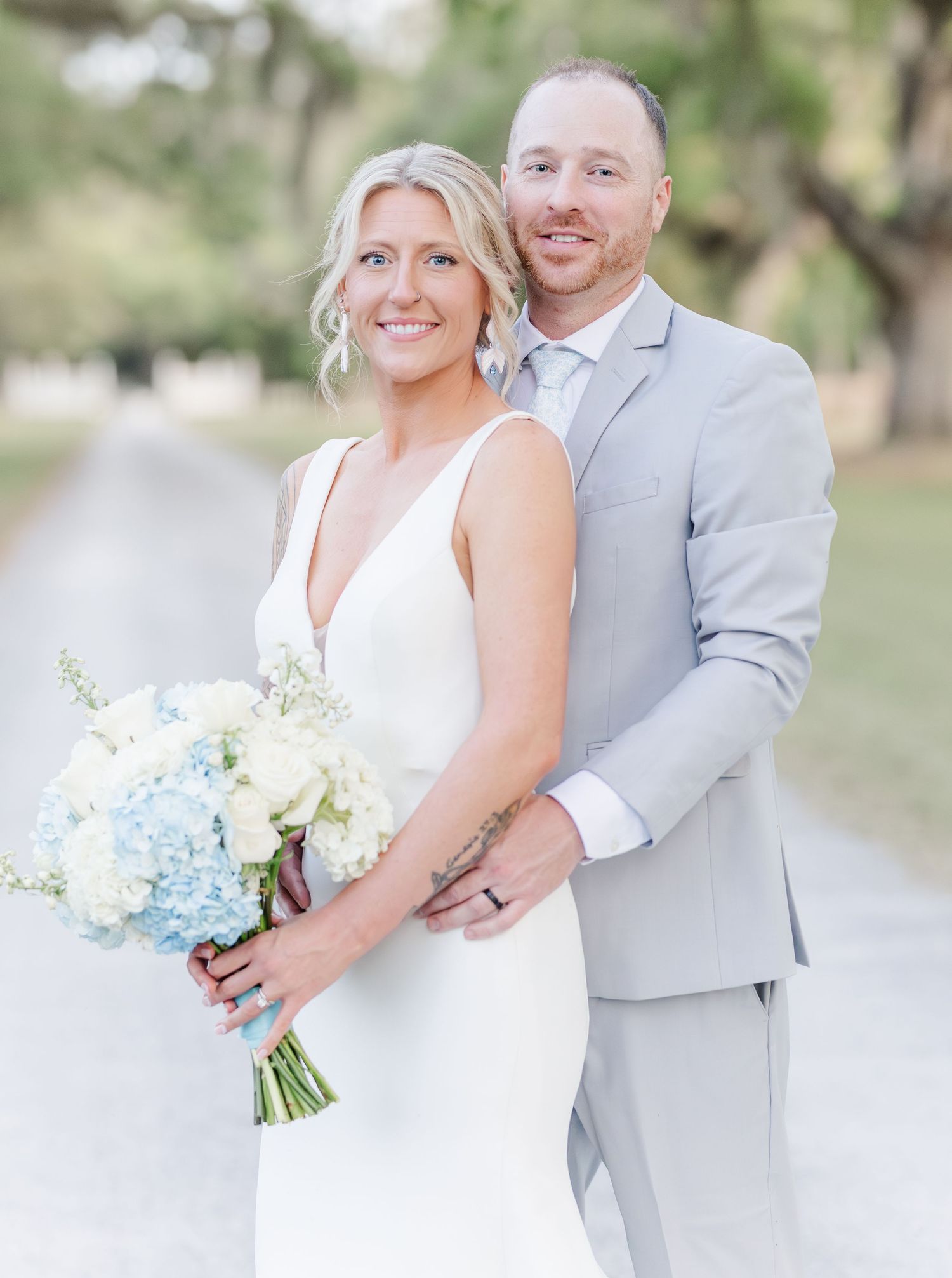 Bride holding blue and white hydrangea bouquet poses with groom on tree-lined path.