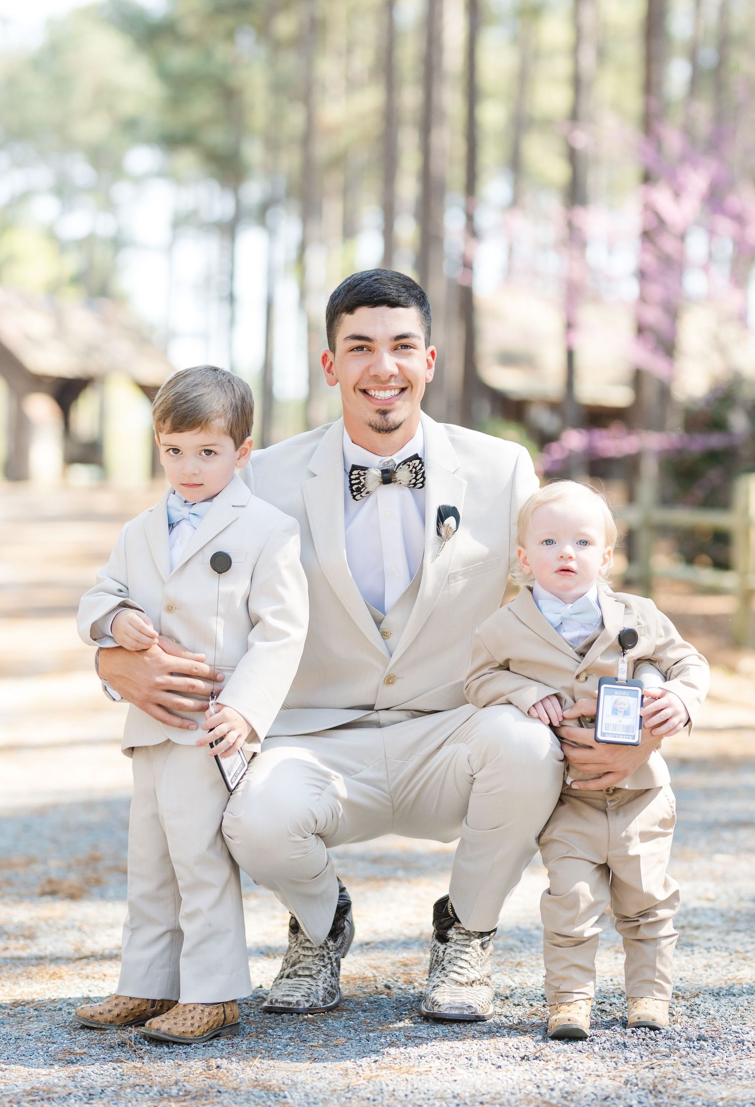 Groom in cream suit crouching with two young ring bearers in matching tan suits at outdoor wedding.