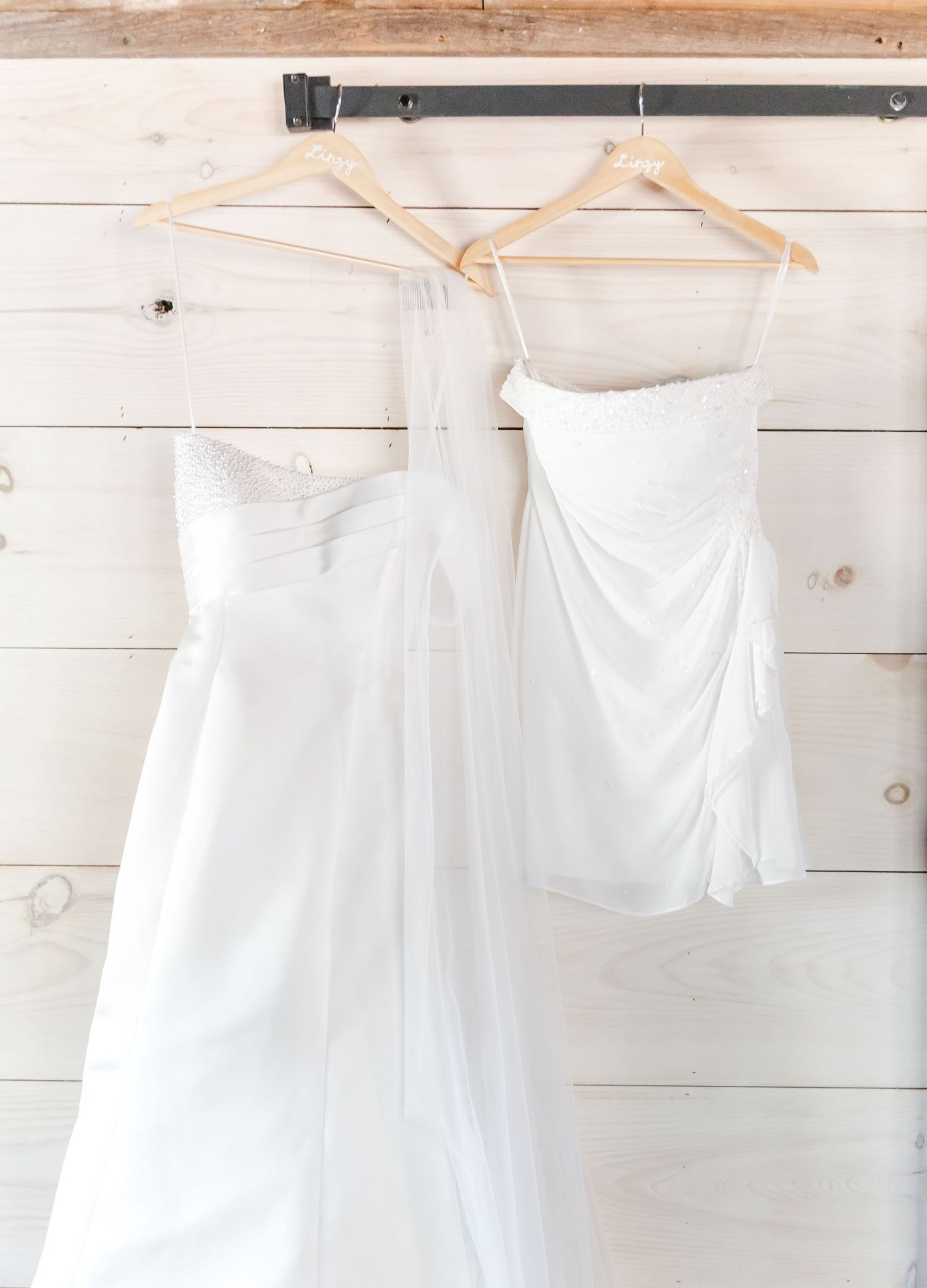 Two white wedding dresses hanging on wooden hangers against a rustic shiplap wall in bridal suite.
