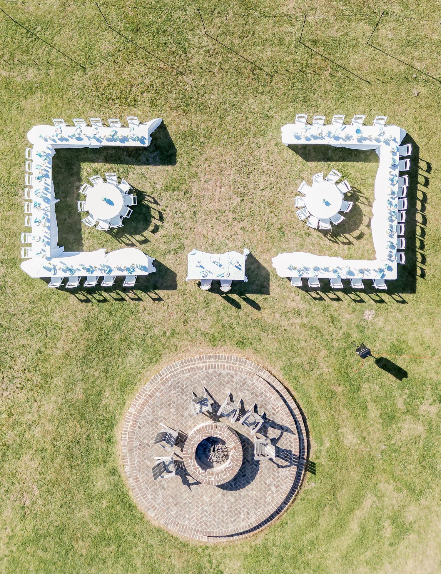 Aerial view of outdoor wedding venue with white chairs, rectangular reception tables, and circular fire pit area.