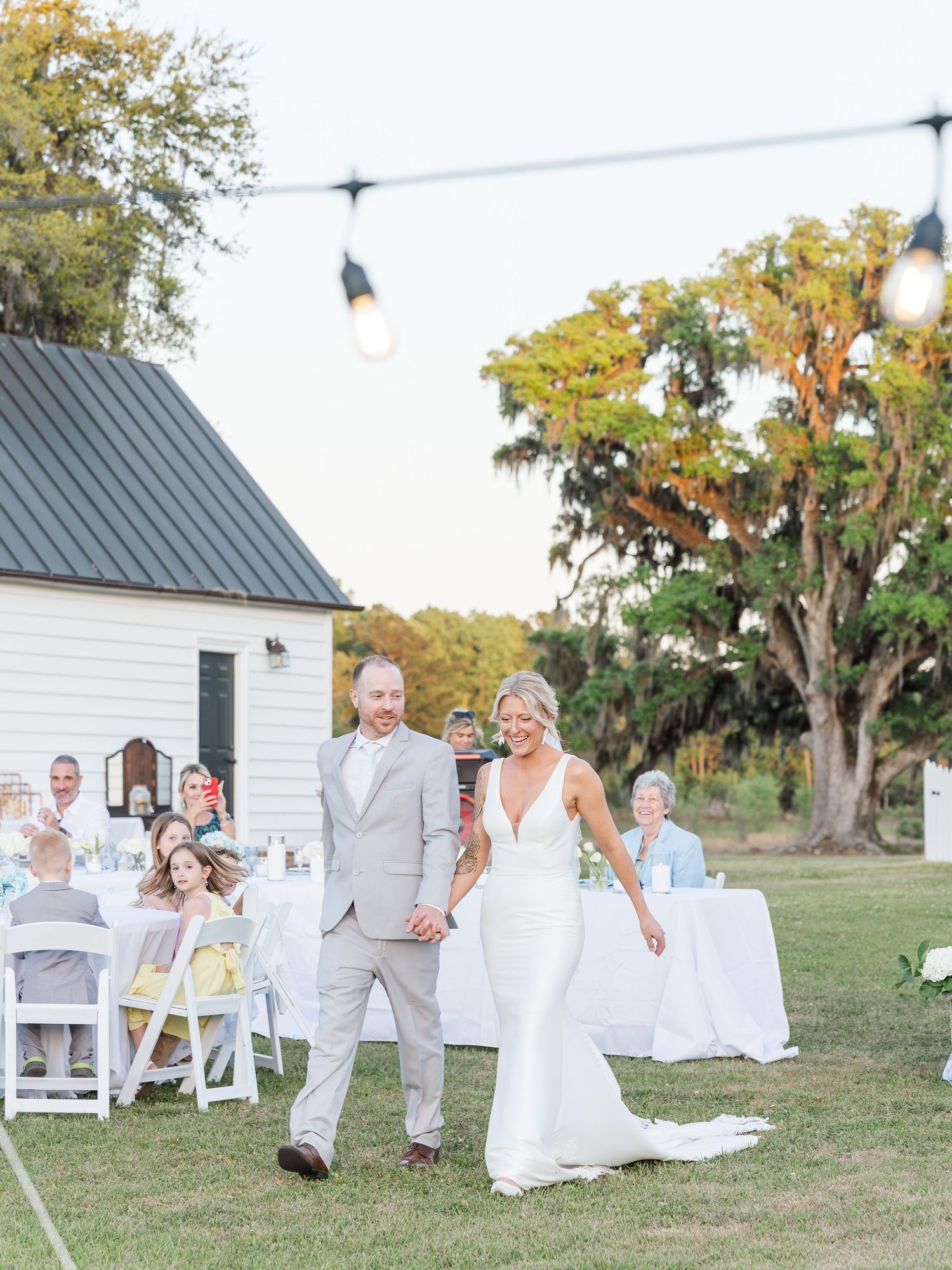 Bride and groom make grand entrance at outdoor evening wedding reception with string lights and white farmhouse.