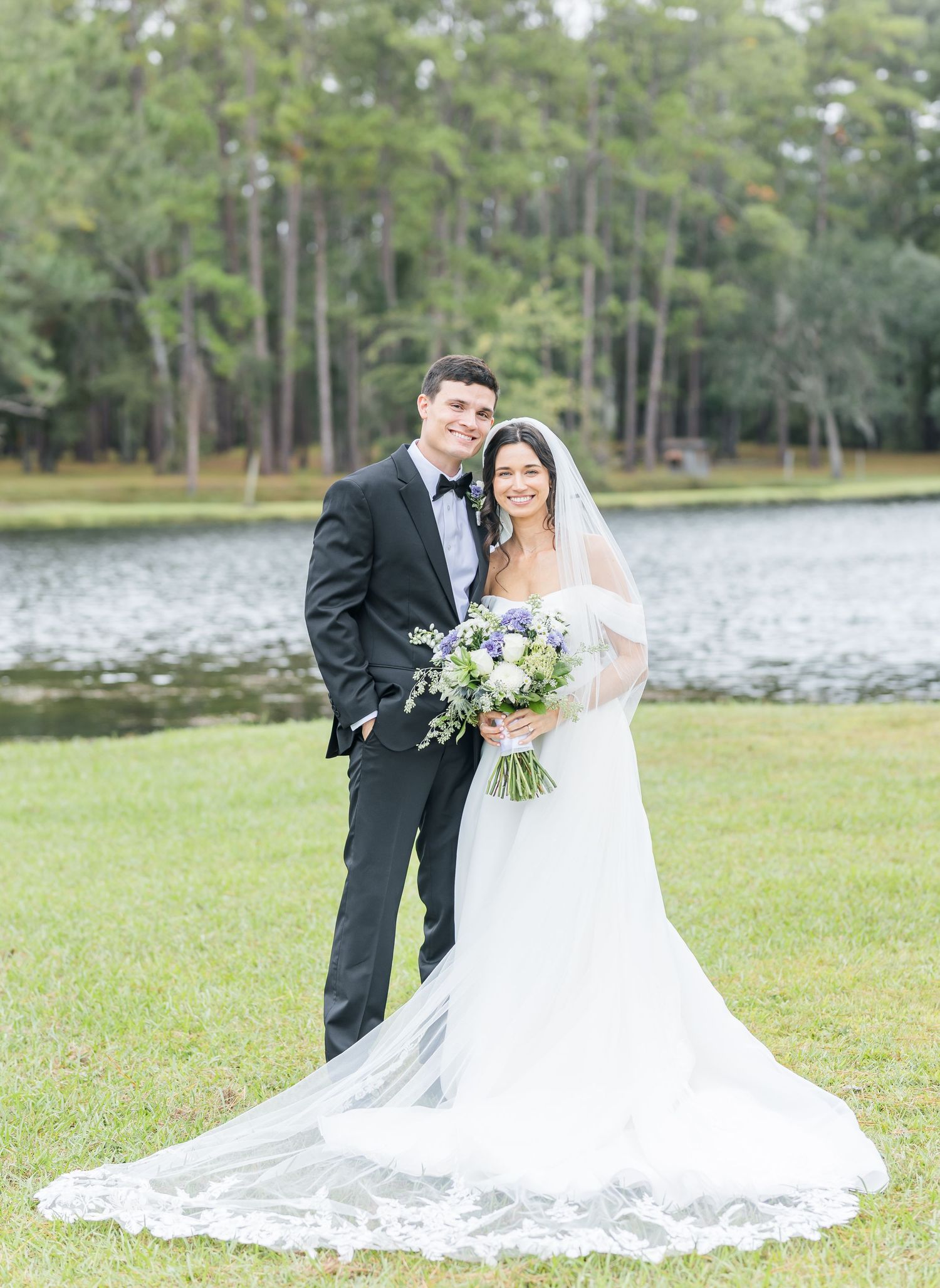 A wedding couple poses together by a serene lake surrounded by pine trees in a romantic outdoor setting.