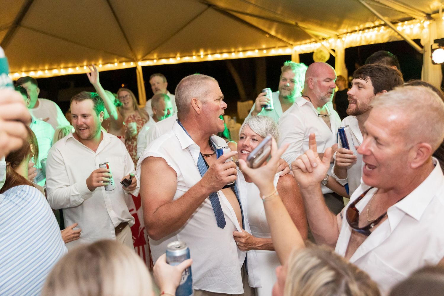 Energetic party crowd celebrates under a tent with drinks and raised hands in white attire.