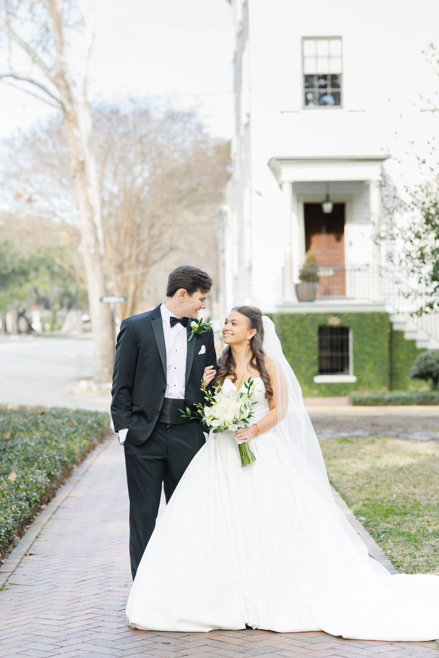 Elegant wedding couple shares romantic moments in formal attire with white bouquet on brick pathway.