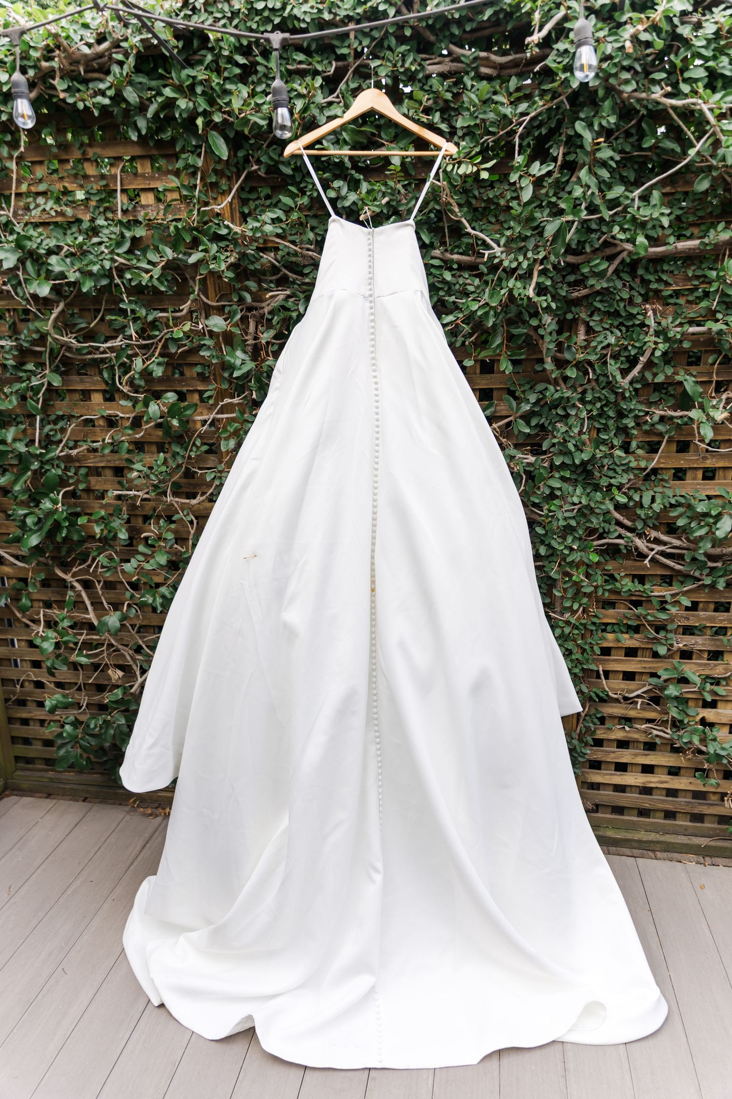 White wedding dress displayed on a hanger against a brick wall with green ivy backdrop.