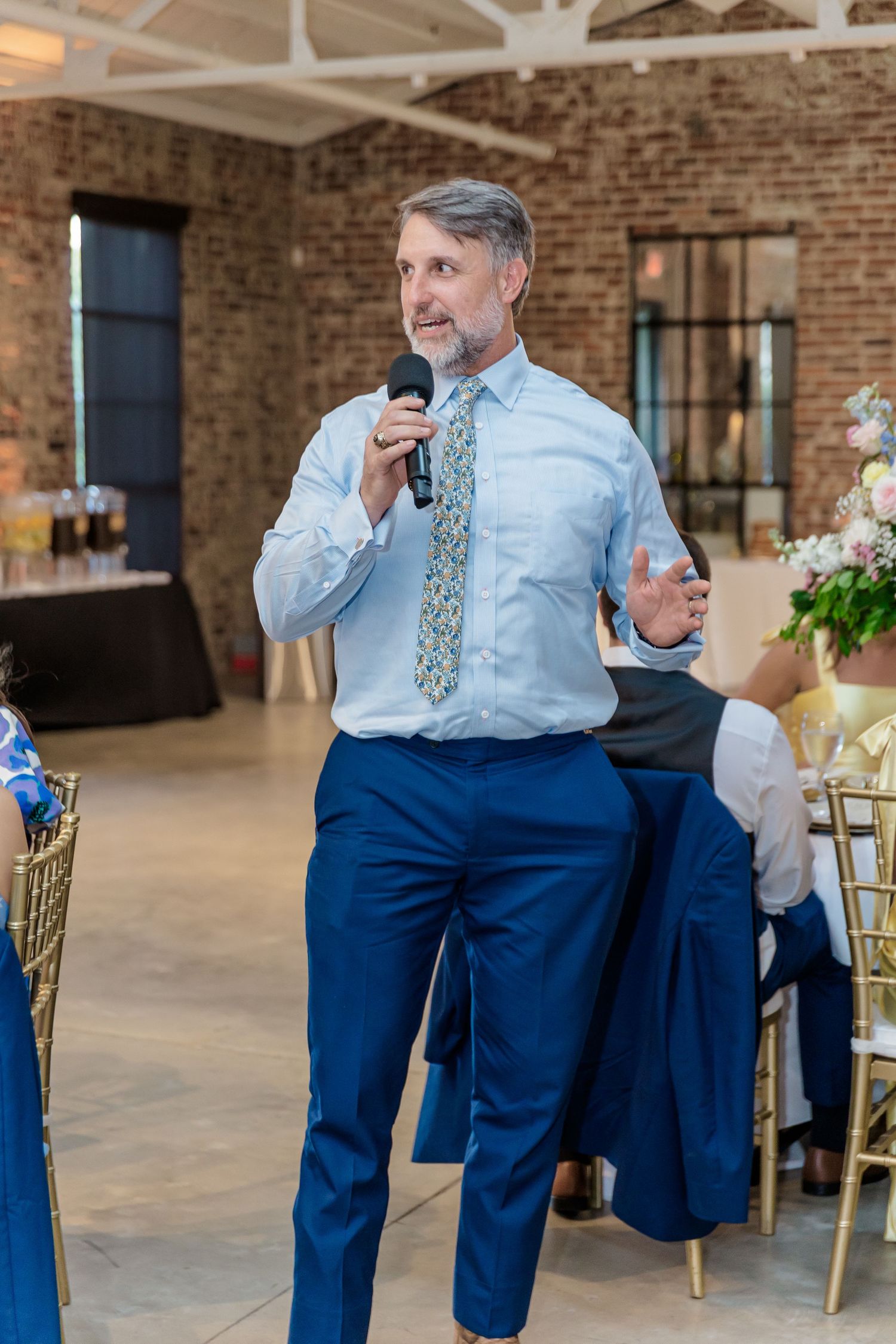 Wedding guests in blue suits and floral ties enjoying a candid moment at an elegant brick-walled reception venue.