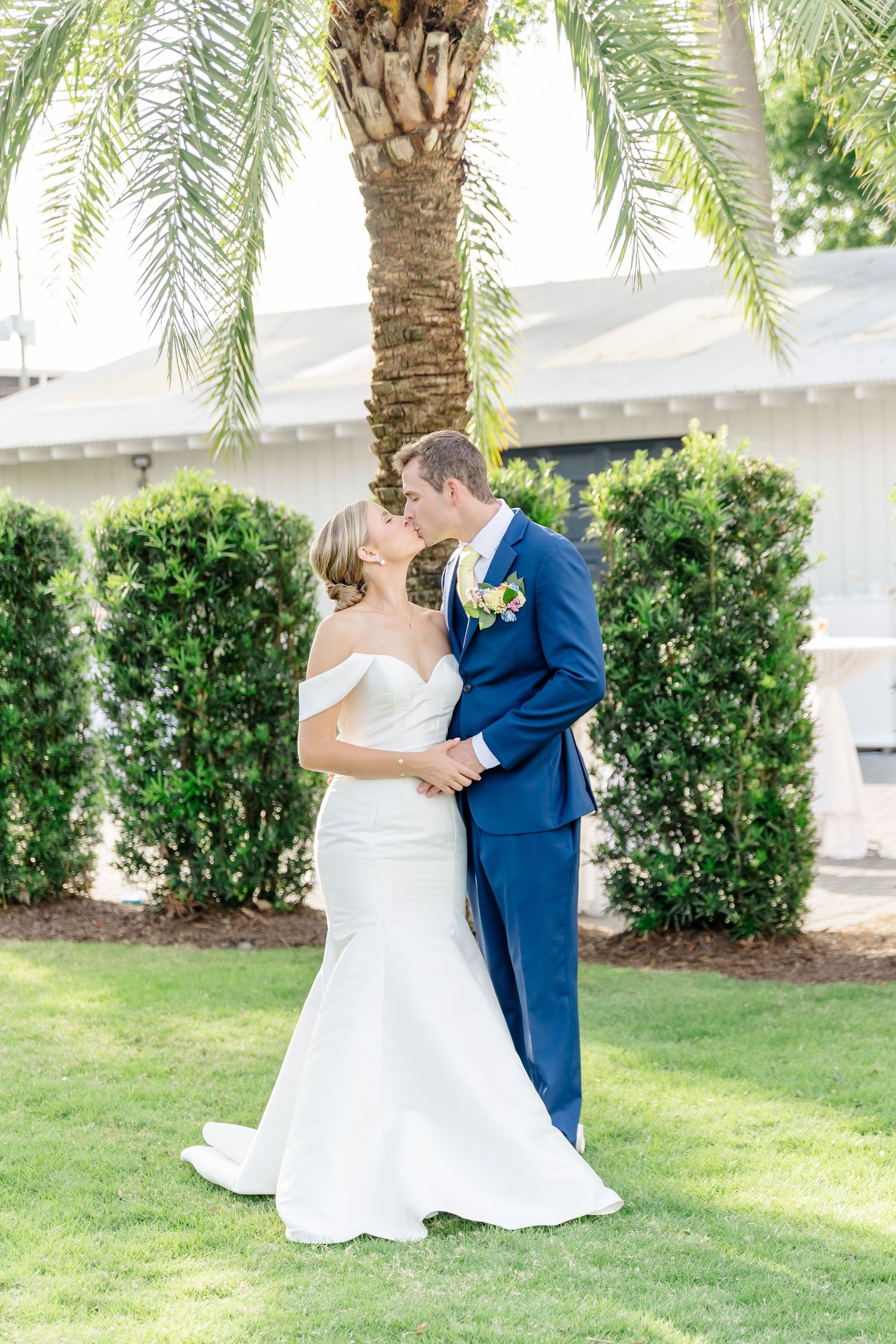 A romantic wedding couple shares intimate moments under palm trees in a garden setting.