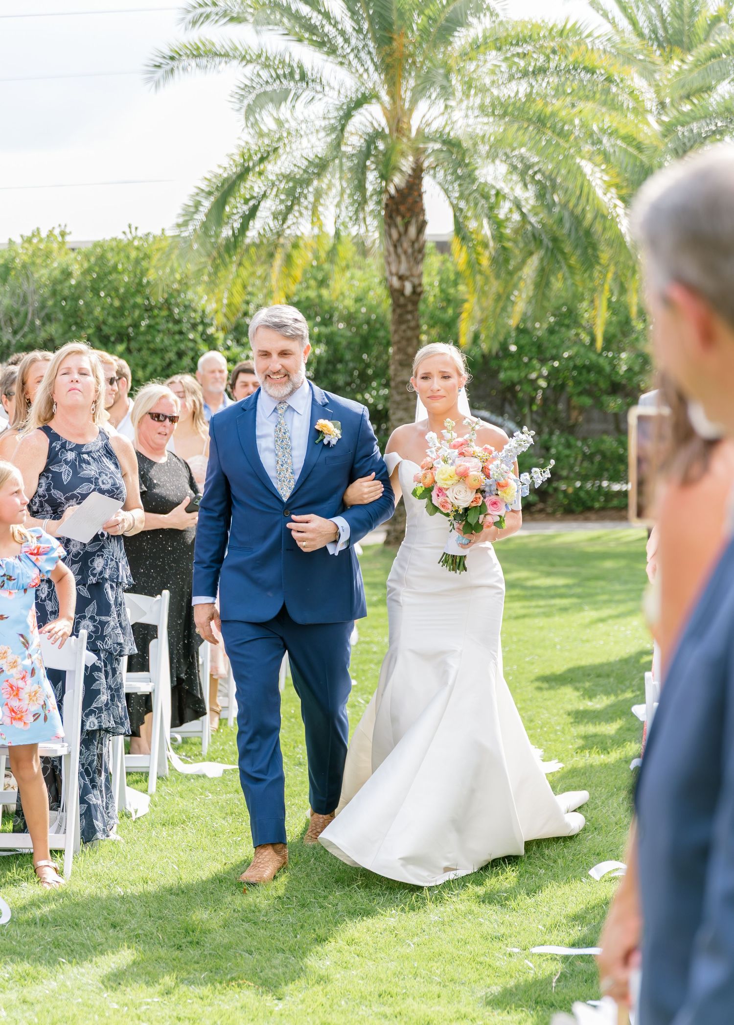 Outdoor wedding ceremony in a lush garden setting with palm trees and guests seated on white chairs.
