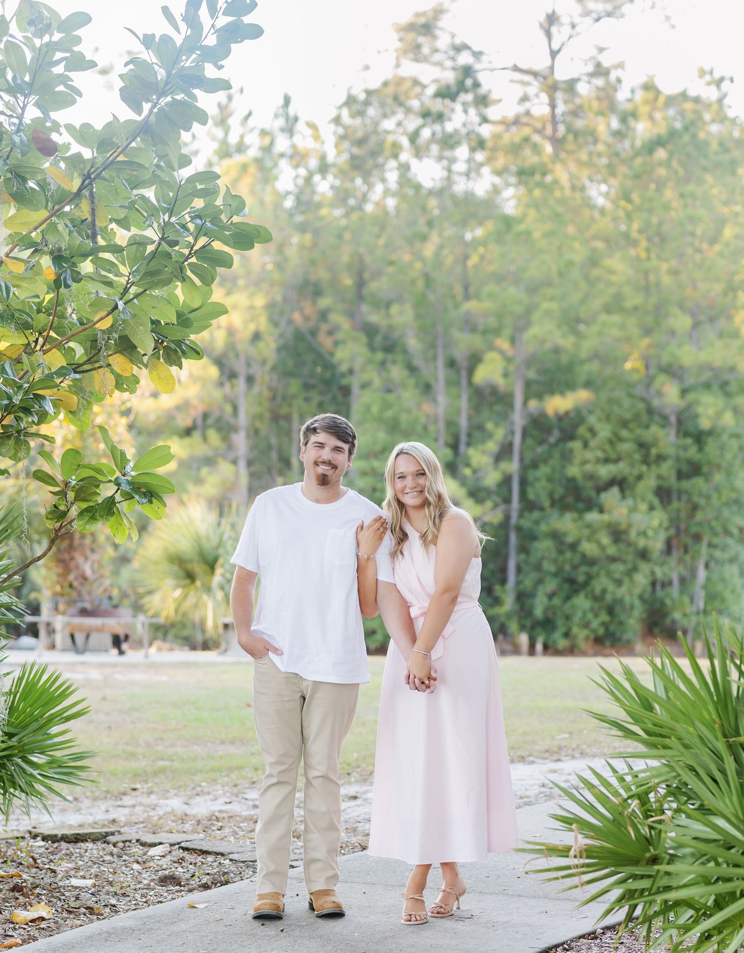 Romantic couple in white and beige outfits walking together in a lush garden setting during golden hour.