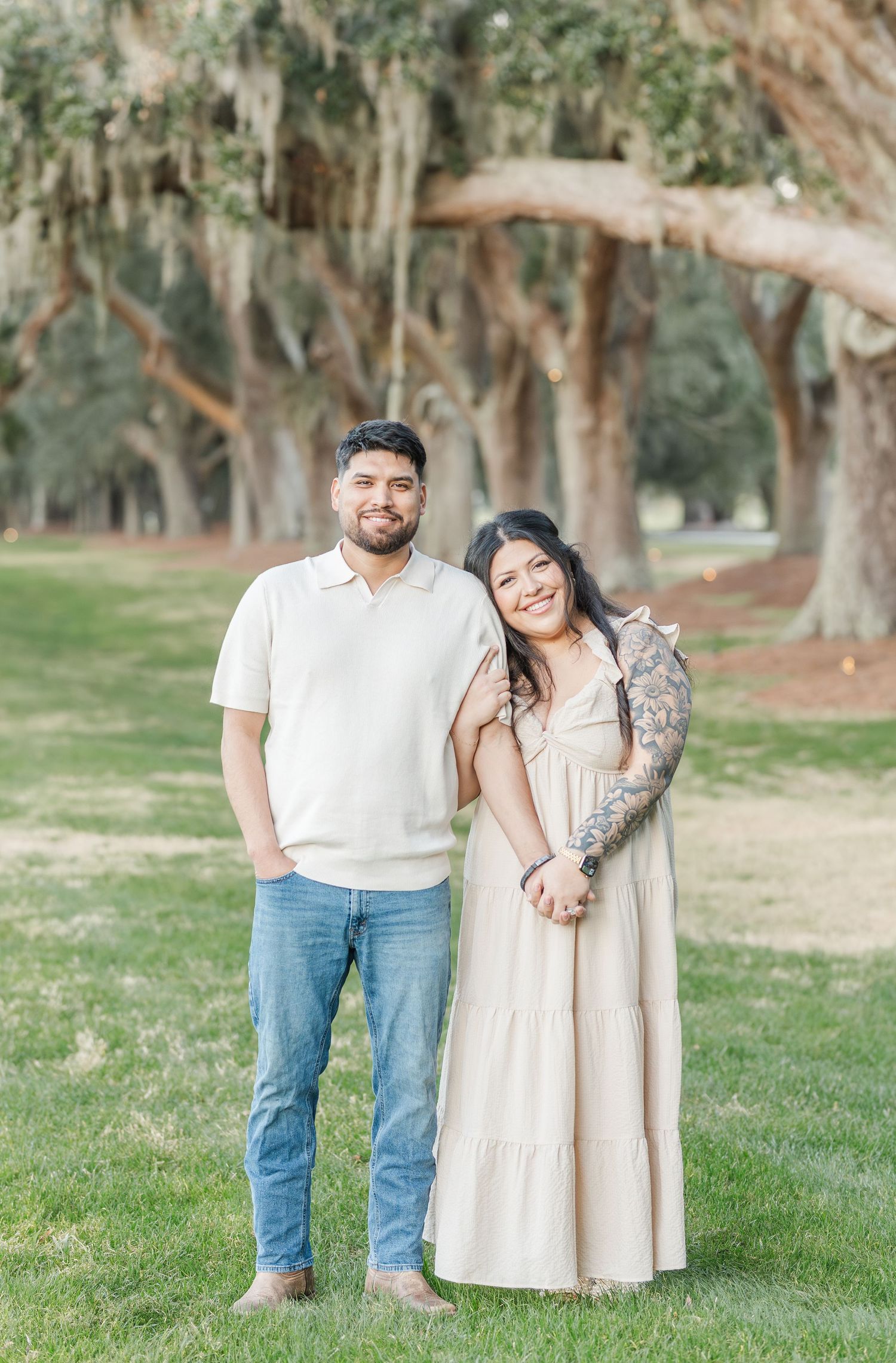 Couple poses together outdoors under Spanish moss-draped trees wearing coordinated cream and white outfits.