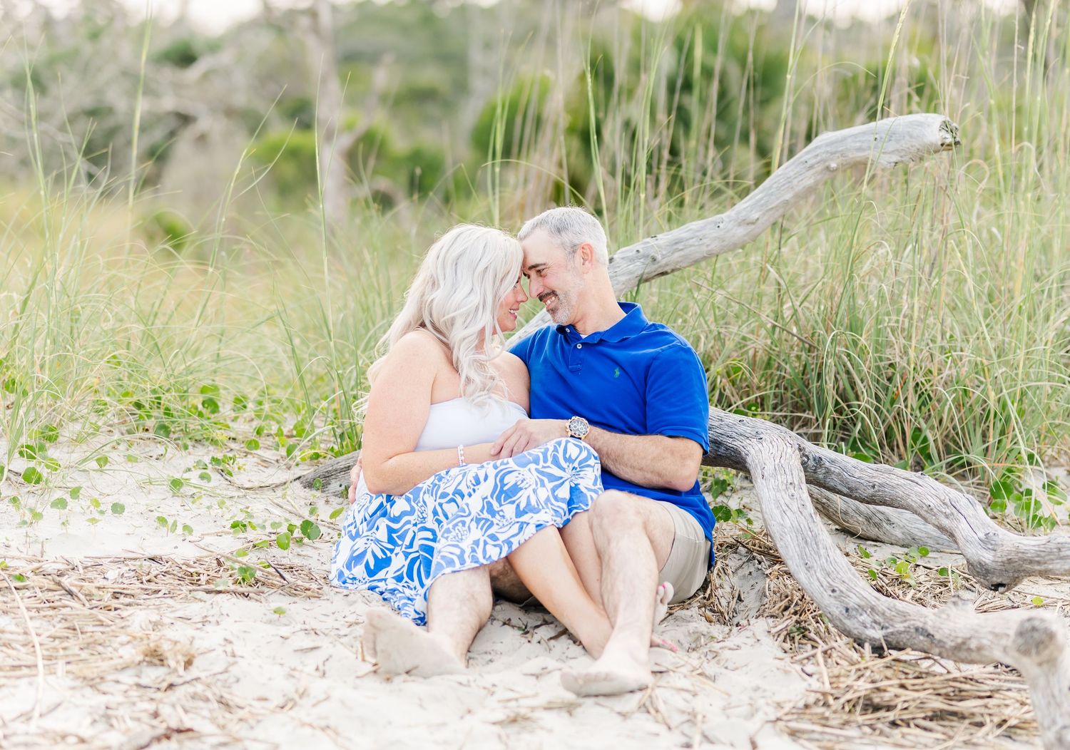 A figure in a blue top and light pants reclines on driftwood at the beach against a soft natural background.