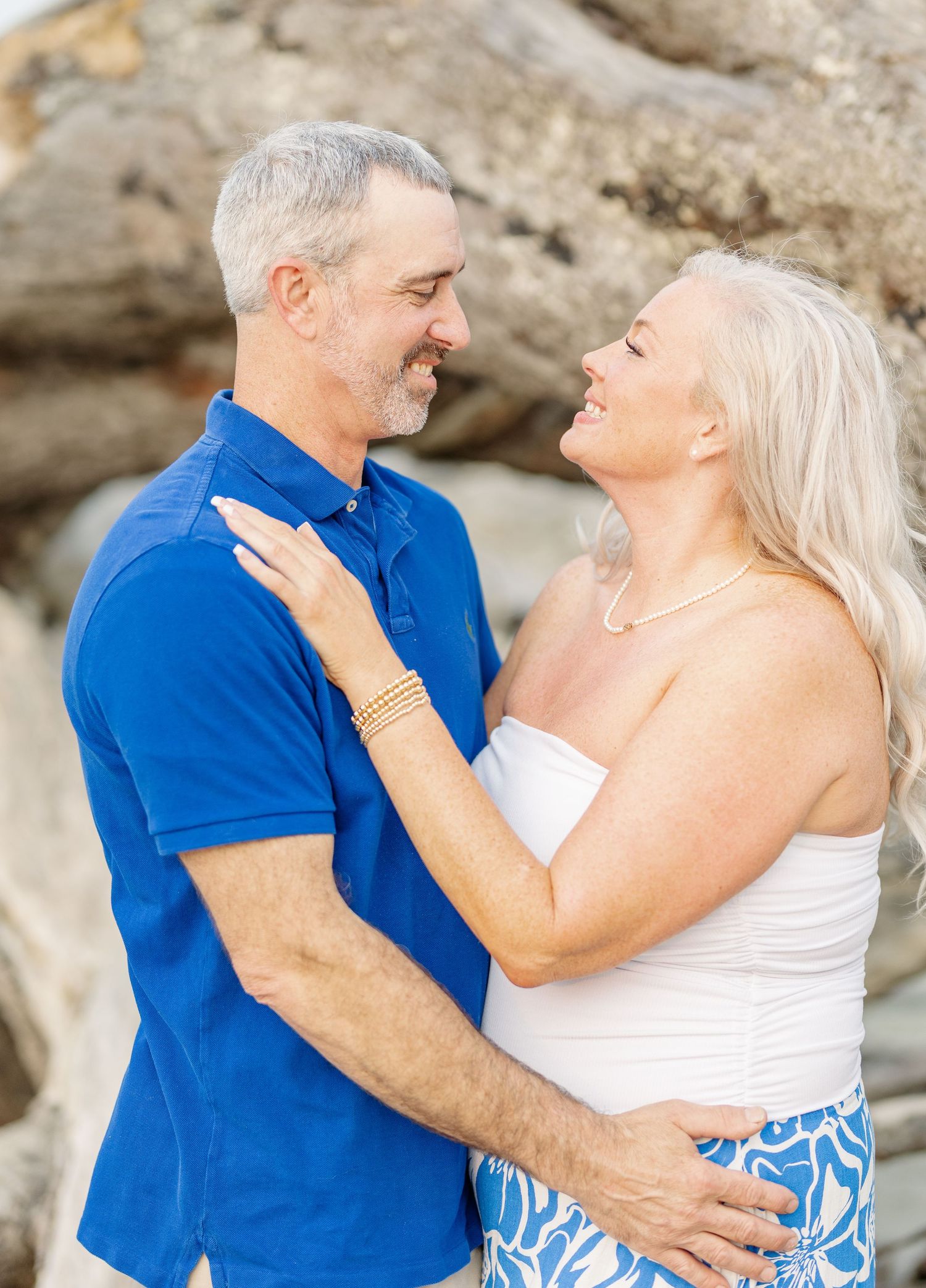 A couple in blue and white outfits share romantic moments against a rocky backdrop at the beach.