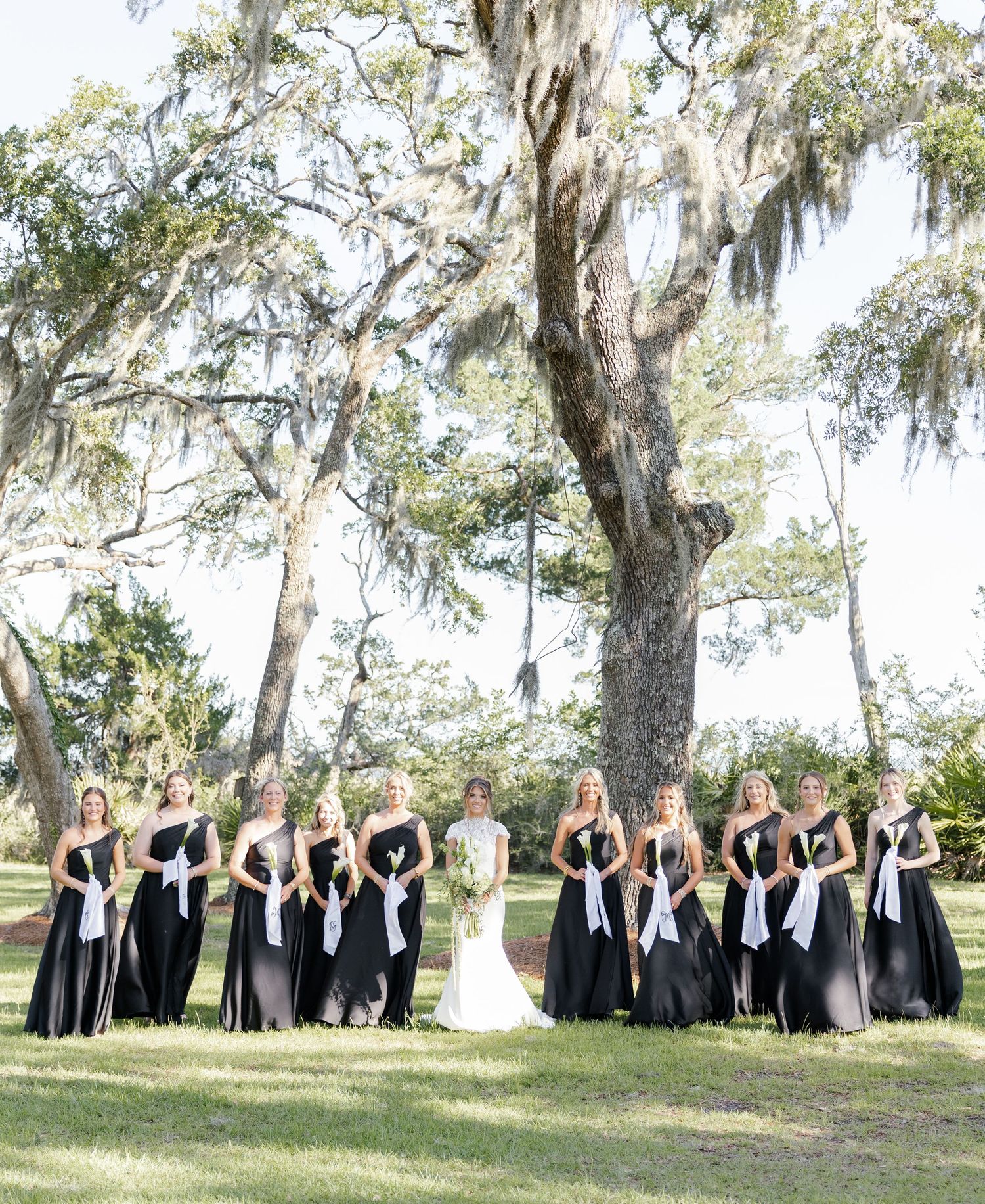 Bride with bridesmaids in black gowns with white sashes posing under Spanish moss-draped oak trees outdoors.