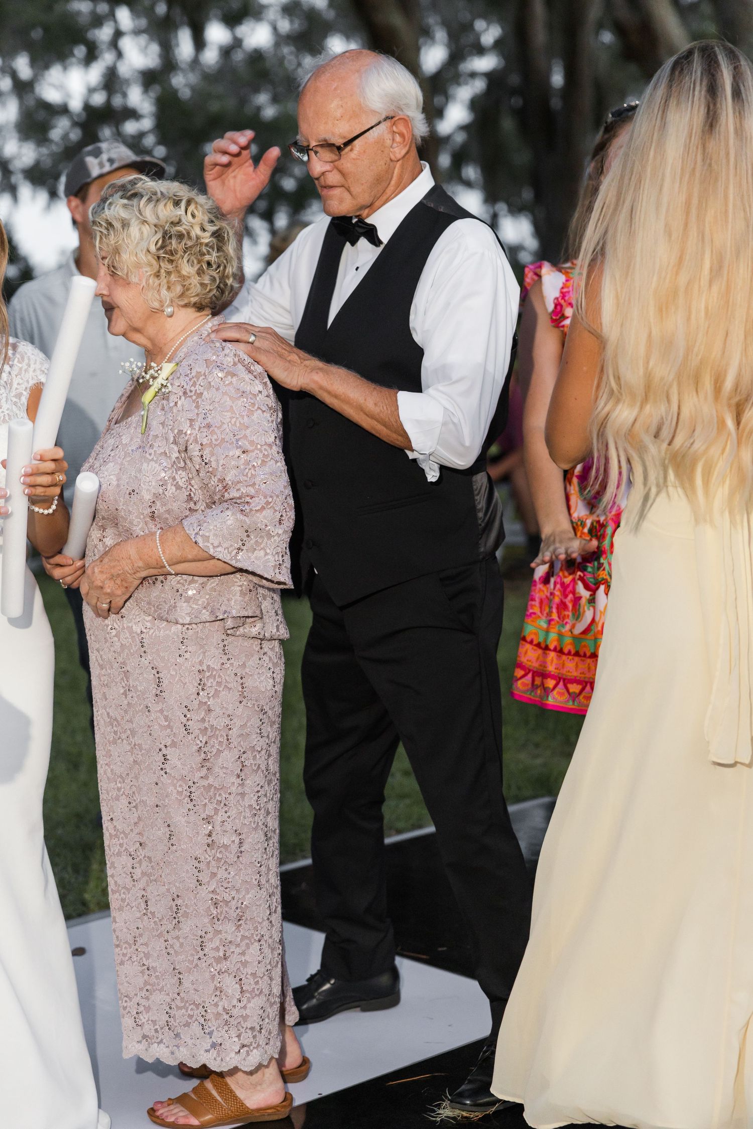 Elderly couple in formal attire walking together at an outdoor wedding ceremony at dusk.