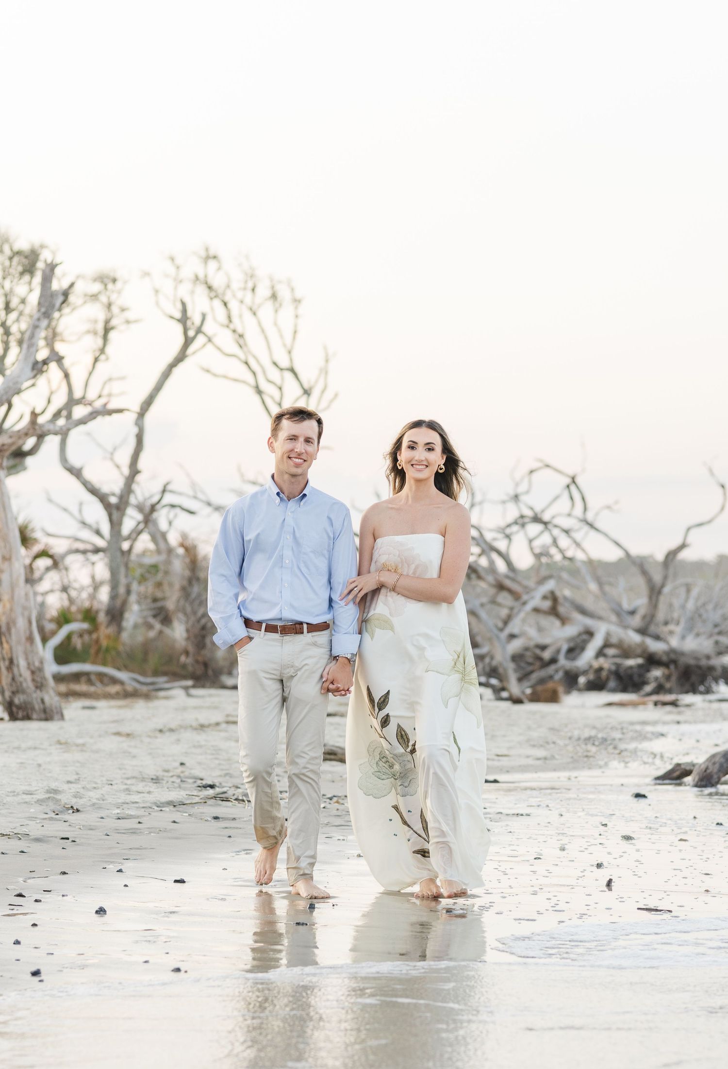 Couple walking hand in hand on beach at sunset with driftwood trees, woman in white floral dress.