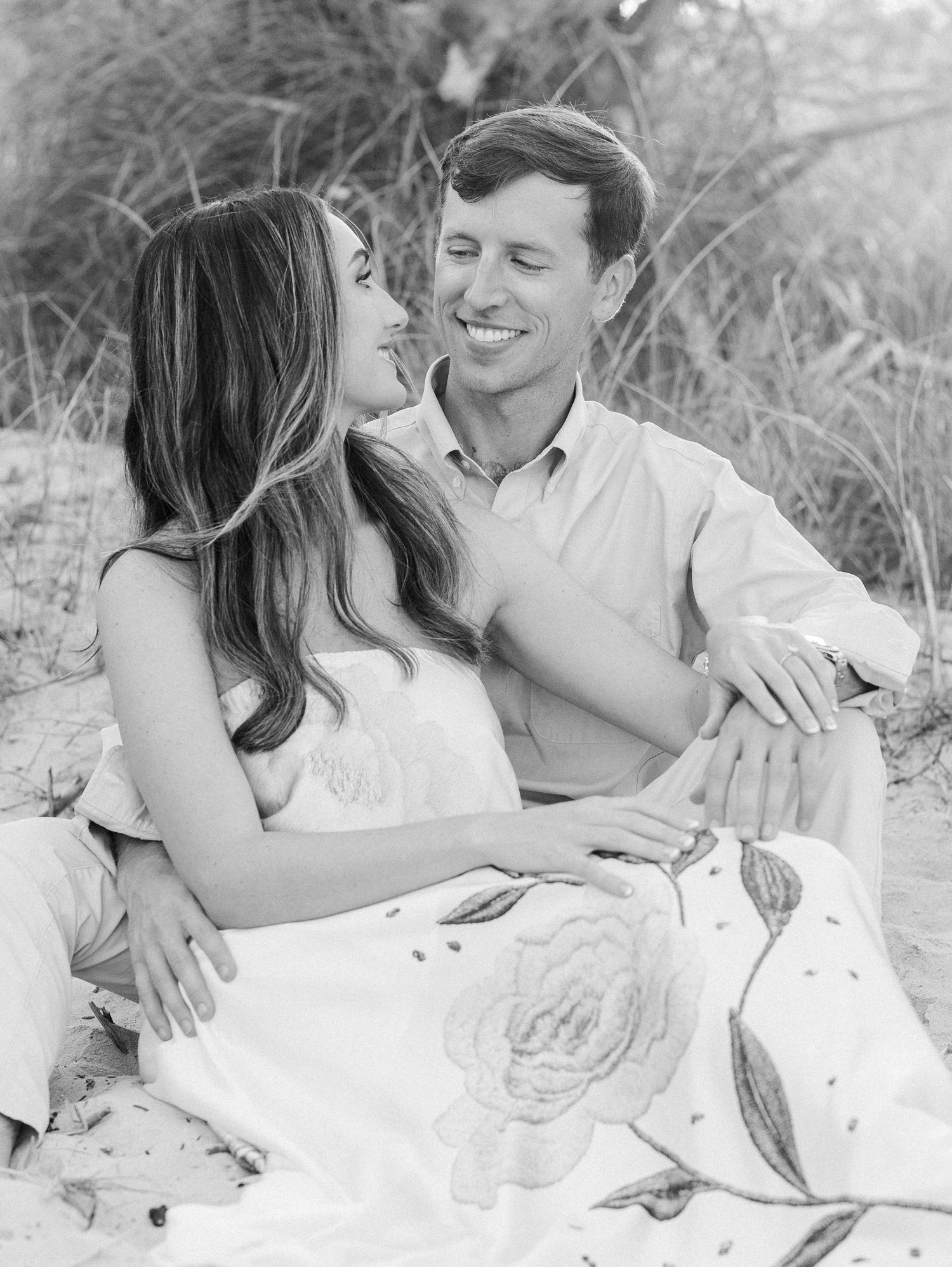 Black and white photo of a smiling couple sitting on beach sand, embracing romantically among tall grasses.