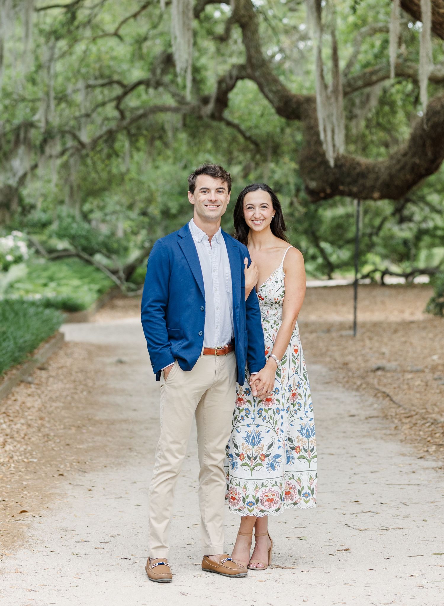 A smiling couple poses on a tree-lined path, man in blue blazer and woman in floral dress, holding hands outdoors.