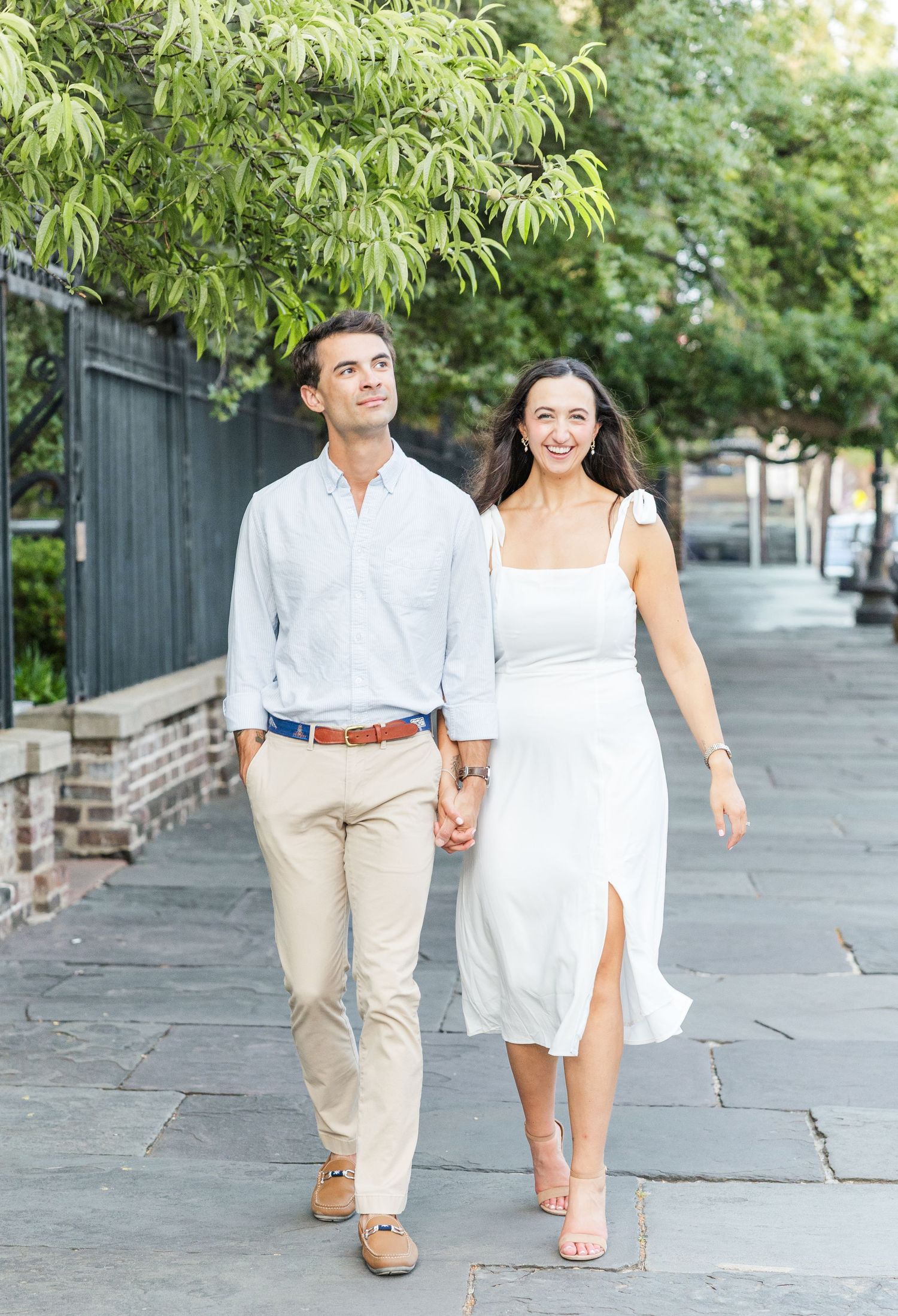 Smiling couple strolling together on a charming stone sidewalk with lush green trees in the background.