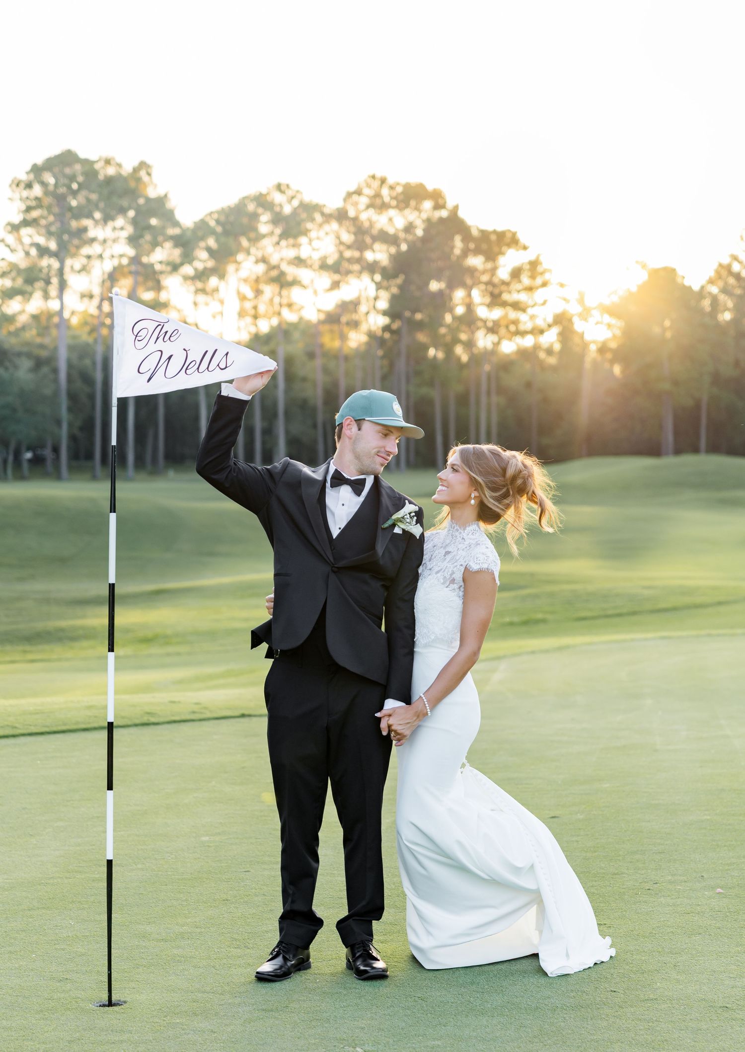 Newlywed couple poses on golf course at sunset, groom holding flag reading The Wells, bride in elegant lace gown.