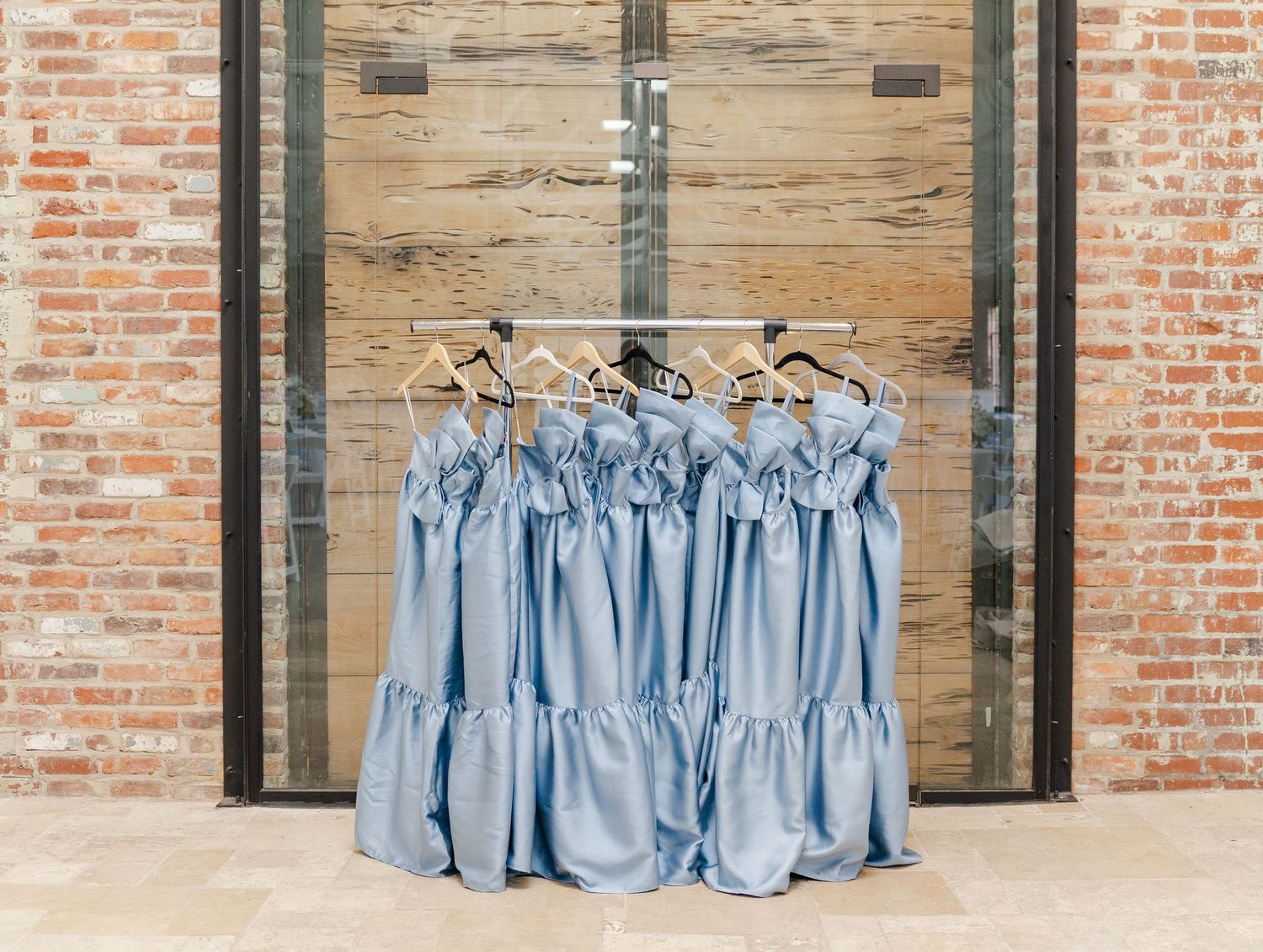 Dusty blue bridesmaid dresses hanging on wooden hangers against a rustic brick wall and glass door.