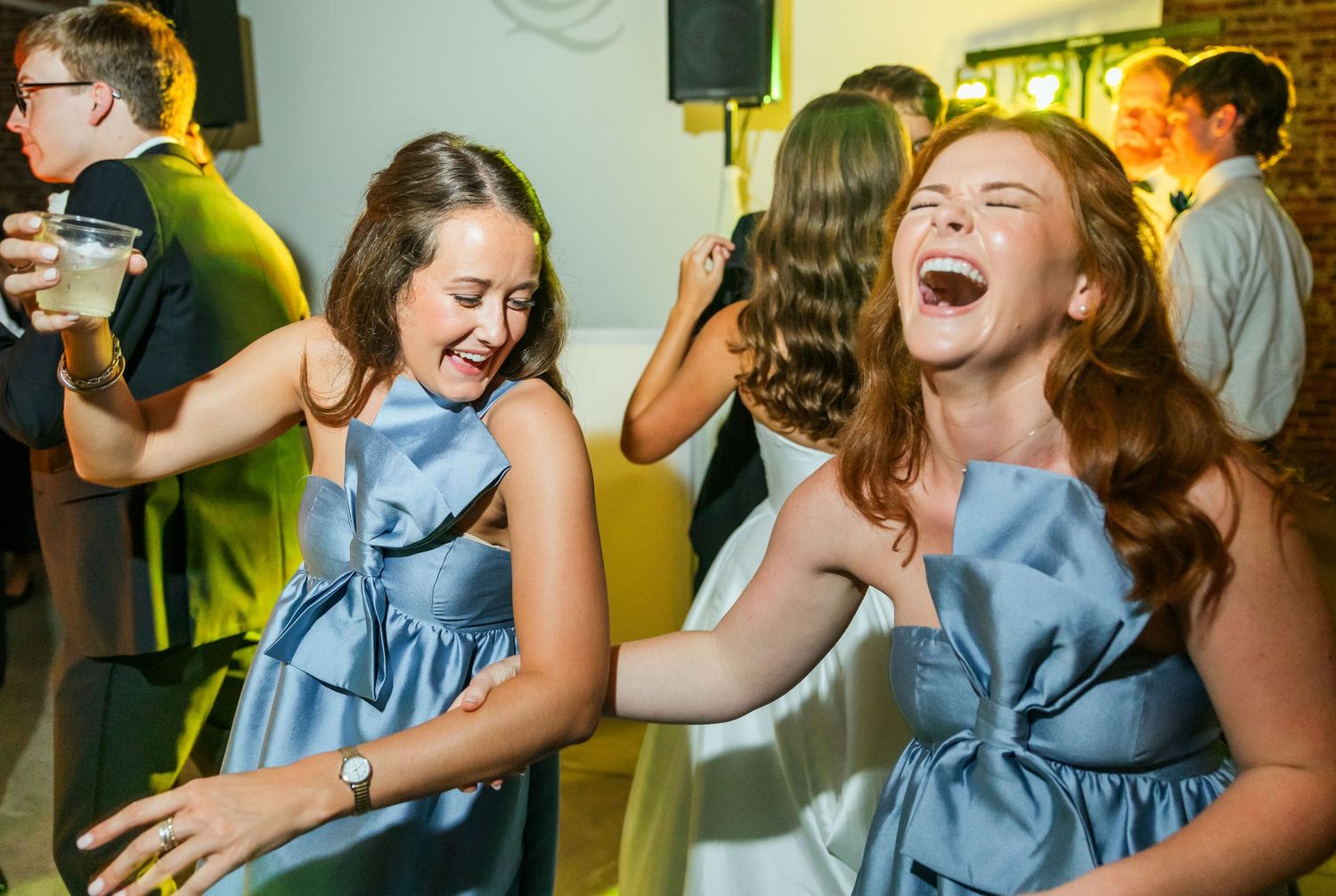 Two bridesmaids in blue dresses laugh and dance together at lively wedding reception.