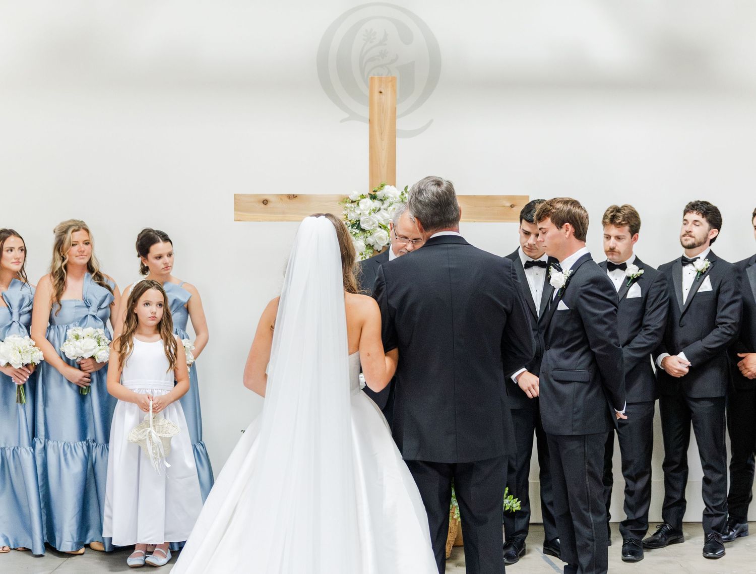 Wedding ceremony at a wooden cross altar with bride, groom, bridesmaids in blue, and groomsmen.