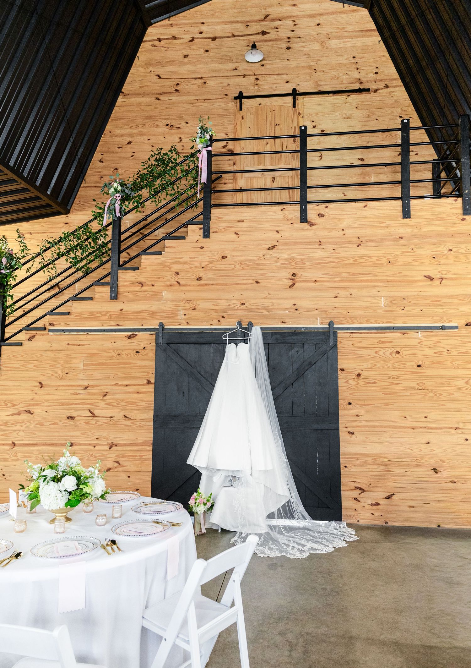 Wedding dress hanging on black barn doors inside a rustic wooden barn venue with reception tables.