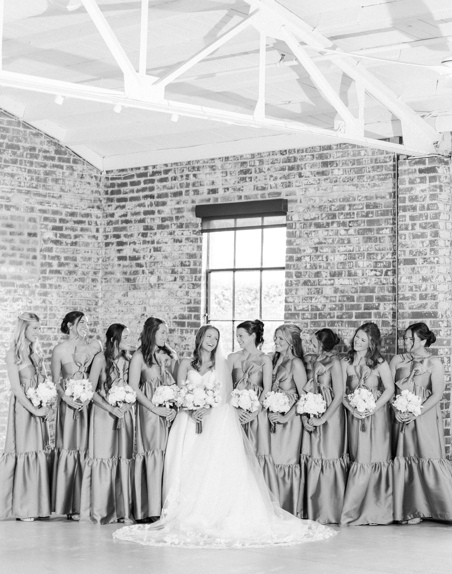Black and white photo of bride surrounded by bridesmaids in matching gowns in brick venue.