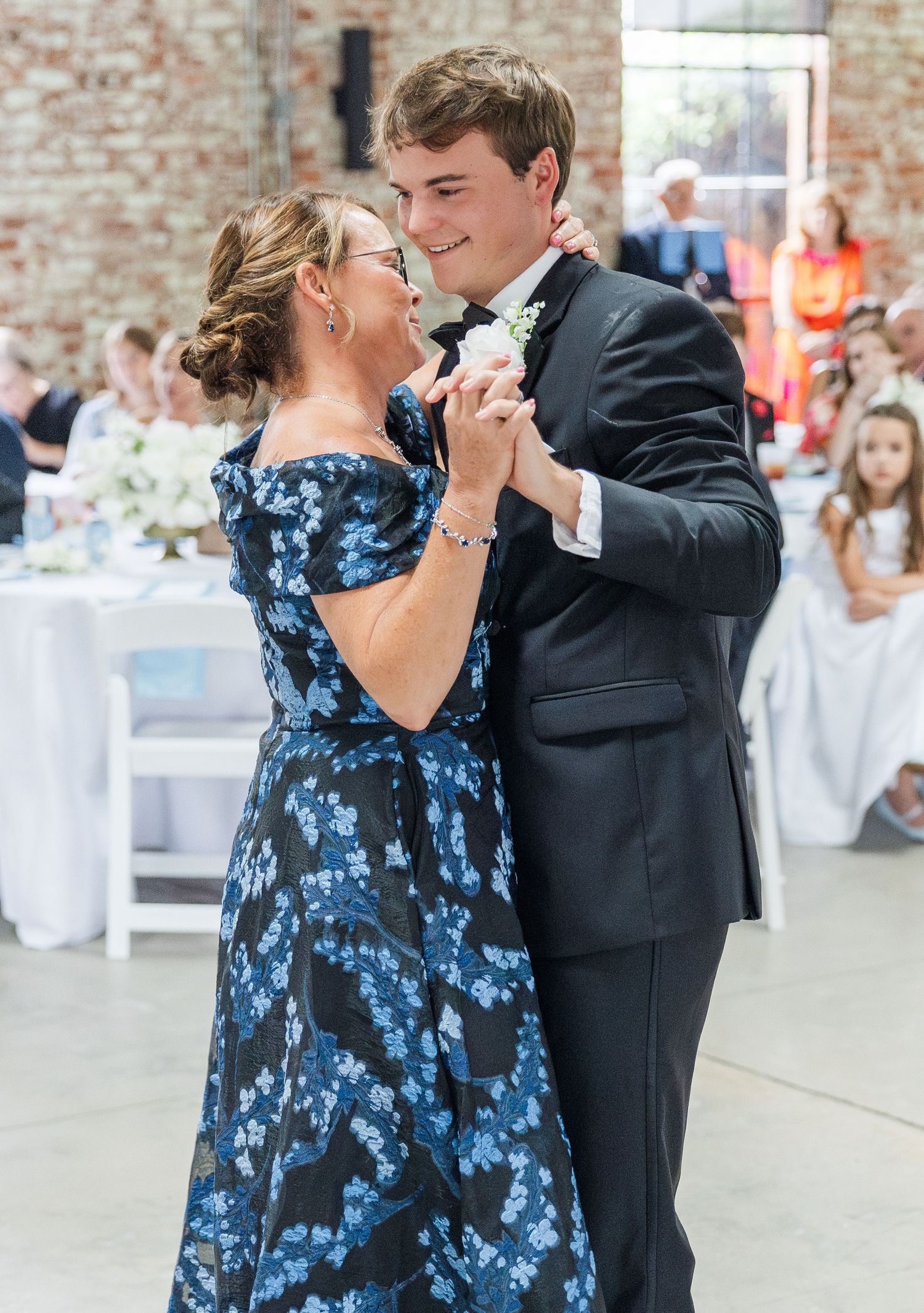 A groom in a black tuxedo shares a tender mother-son dance at an elegant wedding reception.