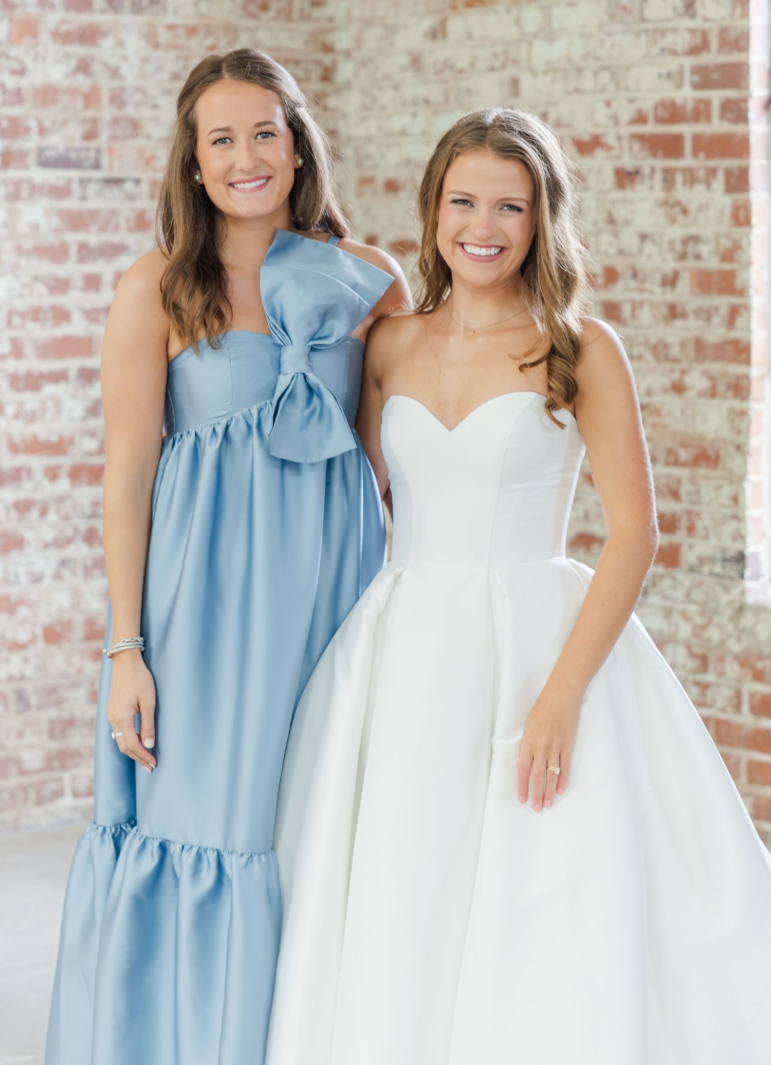 Bride in white gown poses with bridesmaid in blue dress against brick wall.