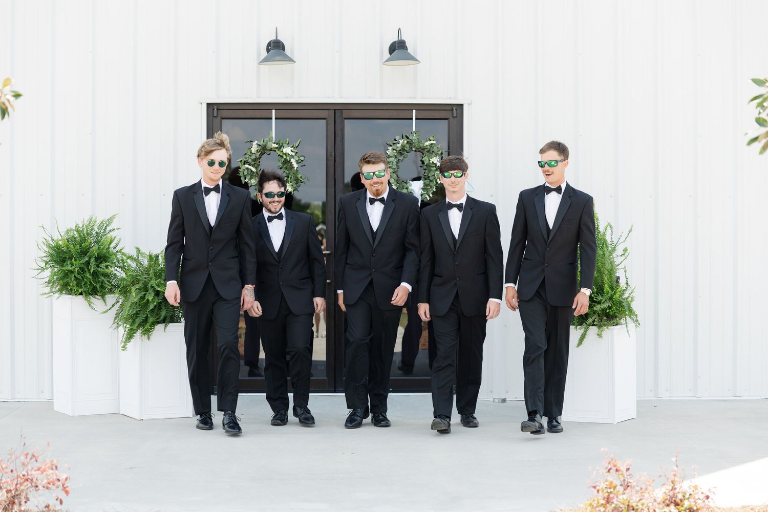 Five groomsmen in black tuxedos and bow ties pose outside a white barn wedding venue.