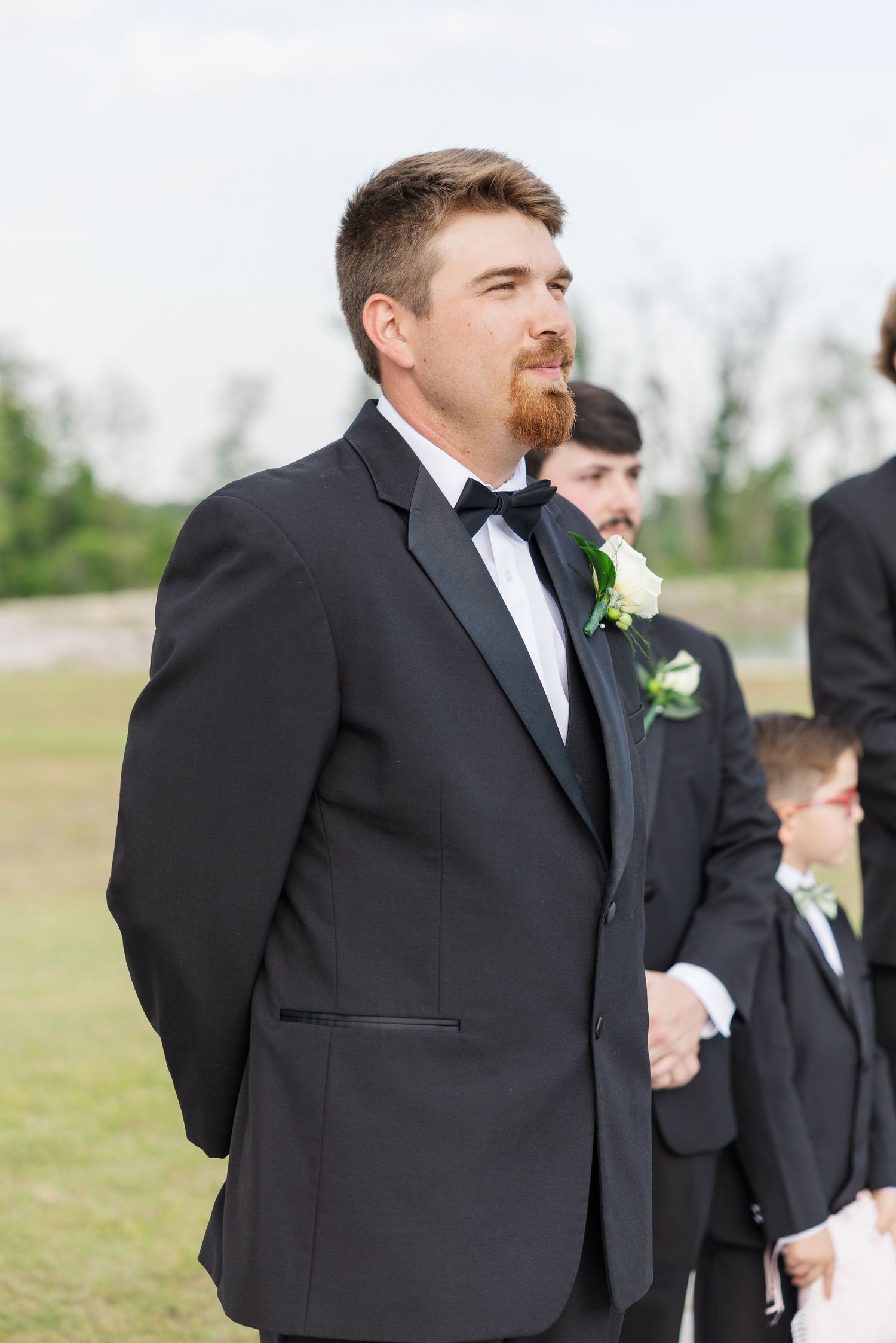 Groom in black tuxedo with white boutonniere stands outdoors with groomsmen during wedding ceremony.