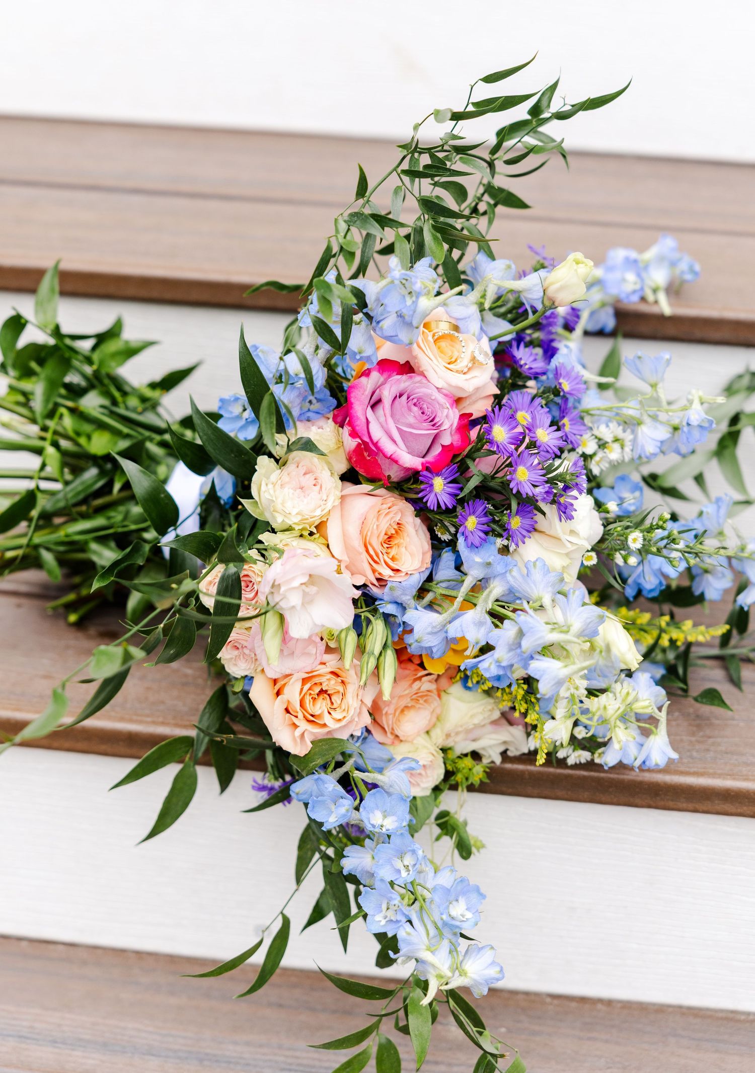 Colorful wedding bouquet with pink roses, blue delphinium, and greenery laid on white wooden steps.