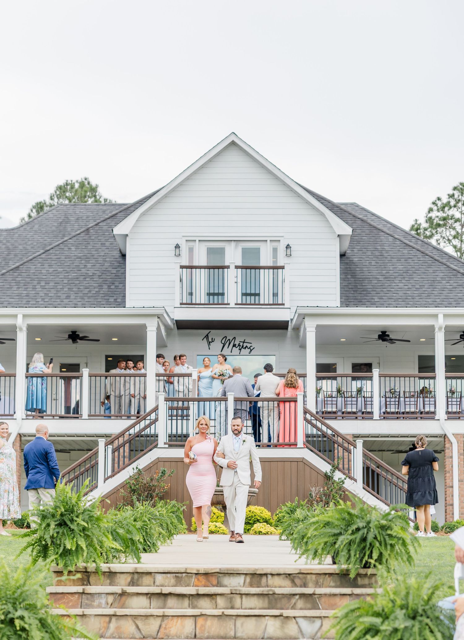 Wedding guests gathered on white farmhouse venue balconies as couple descends stone steps outdoors.