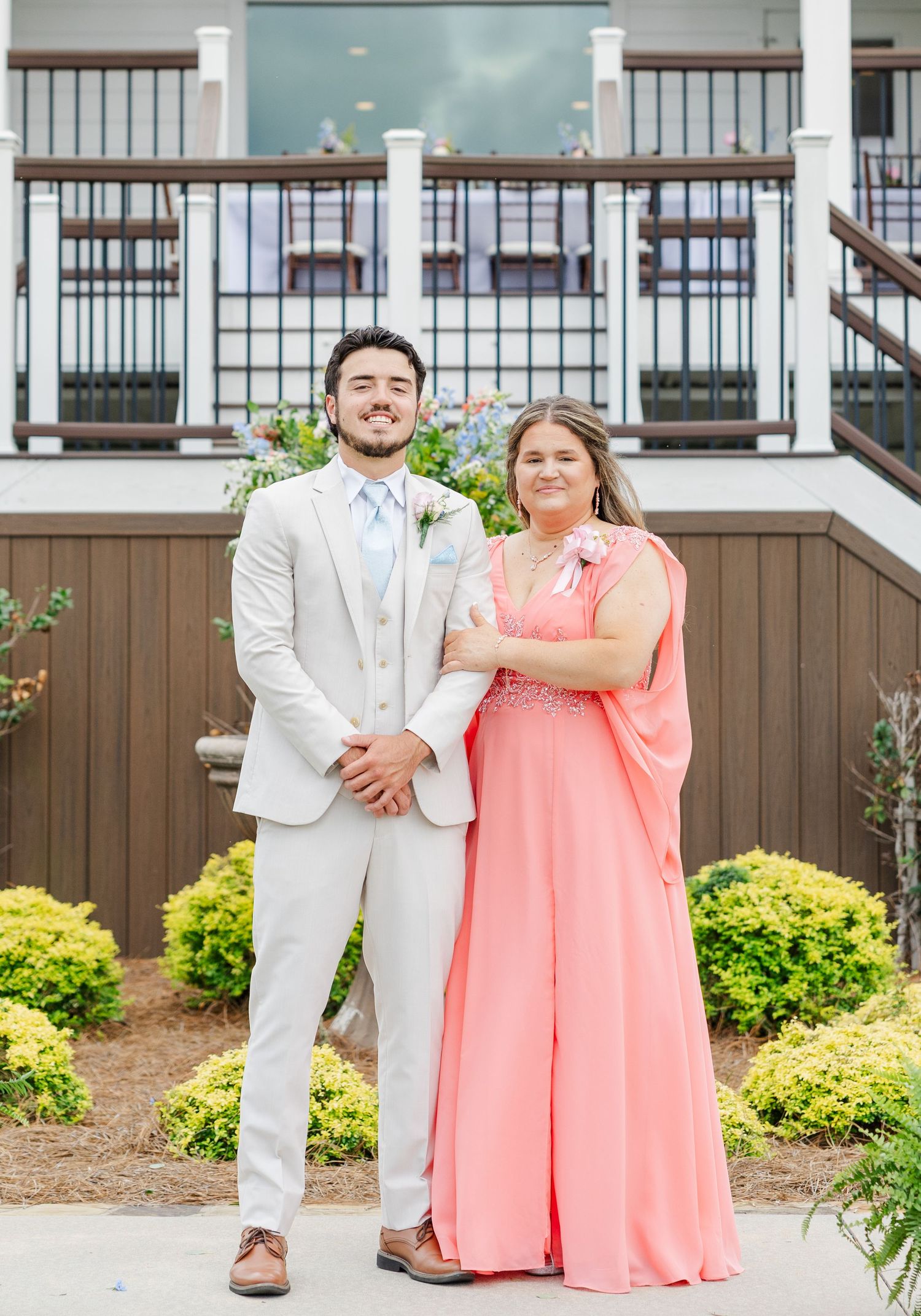 Groom in beige suit stands with woman in coral gown outside elegant wedding venue with decorative balcony.