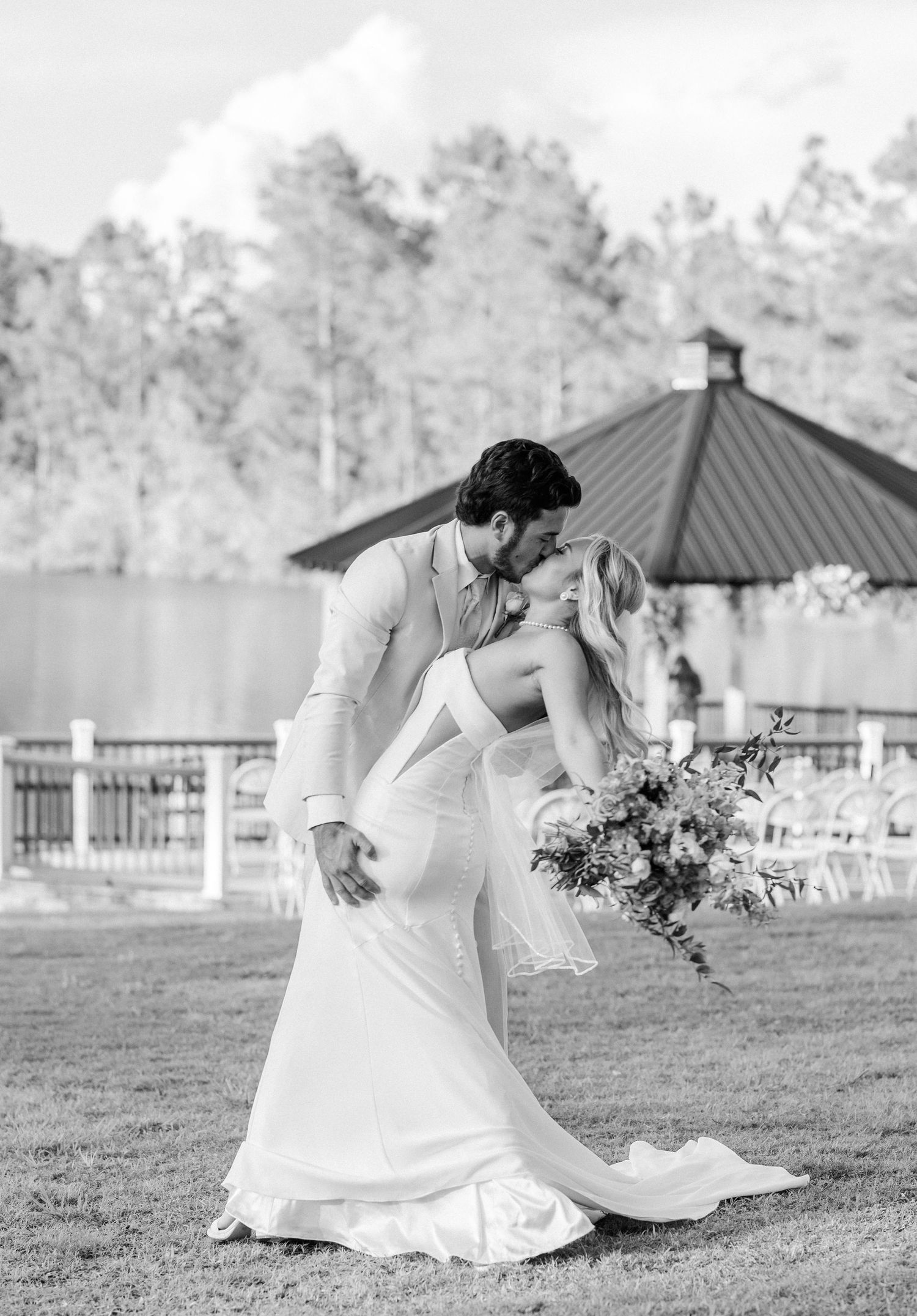 Black and white photo of groom dipping bride for kiss on lawn with gazebo backdrop.