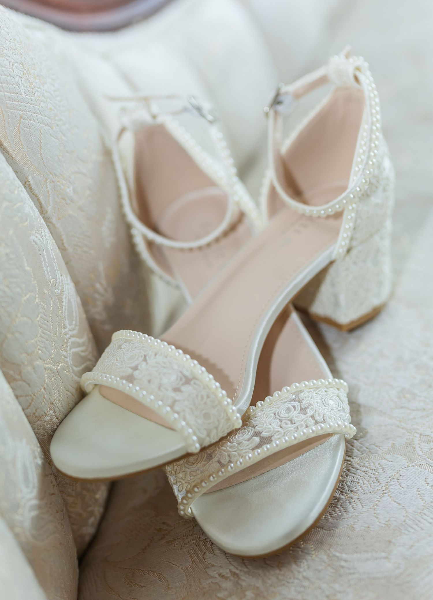 Delicate white bridal shoes with pearl embellishments rest on lace fabric.