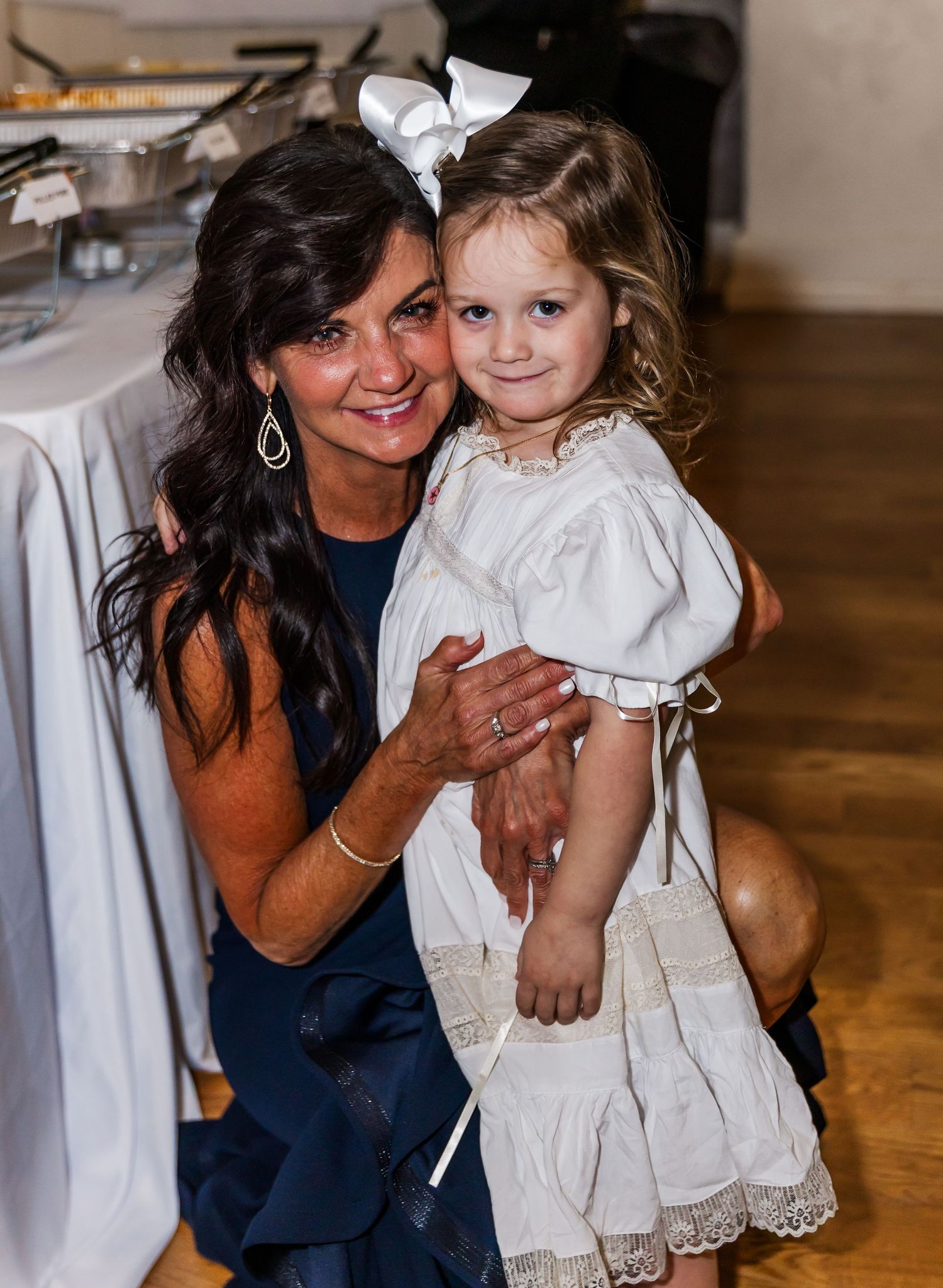 Woman in navy dress smiles holding a young flower girl in white dress with a large bow at wedding.