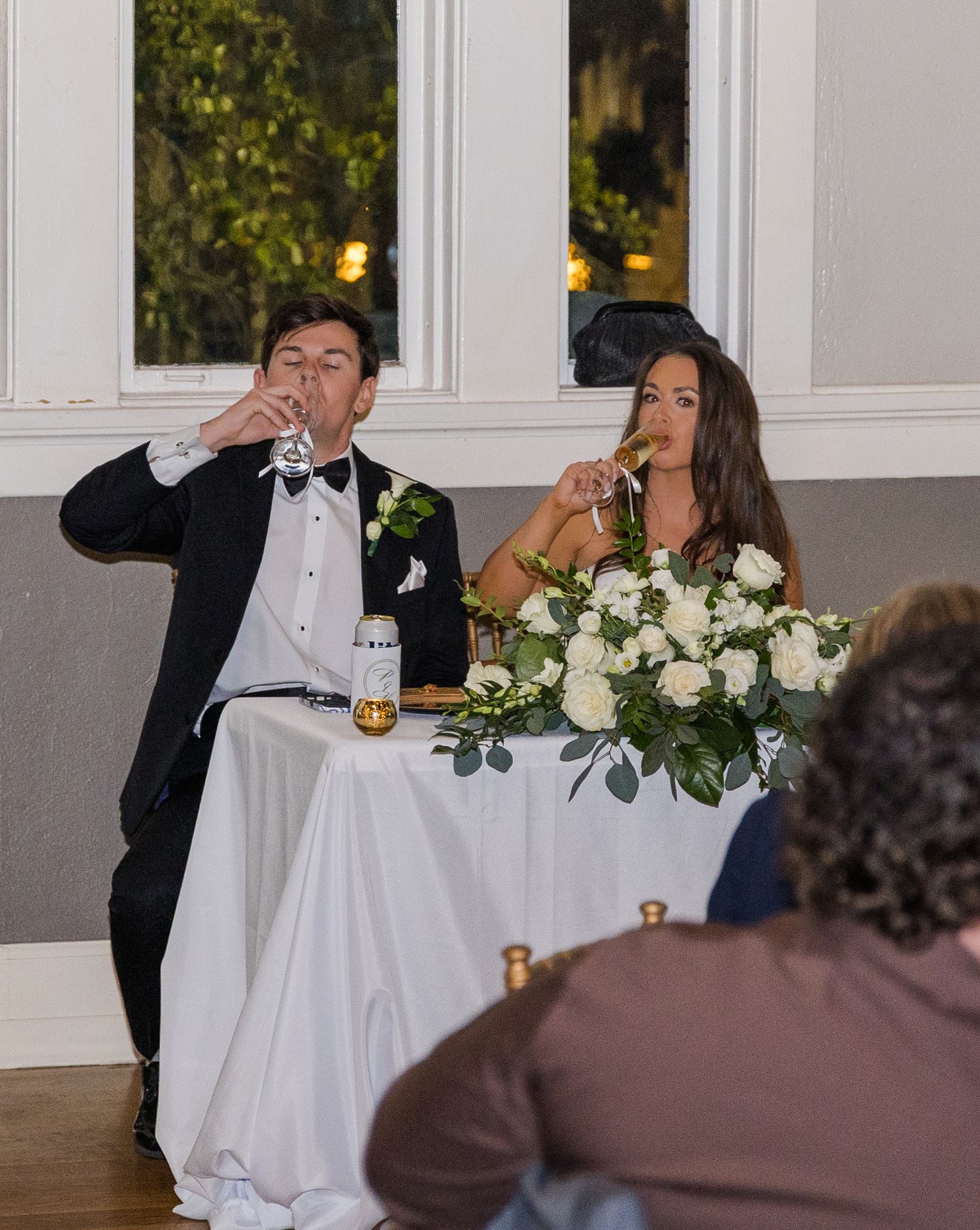 Bride and groom drink champagne toast at sweetheart table with white floral centerpiece at reception.