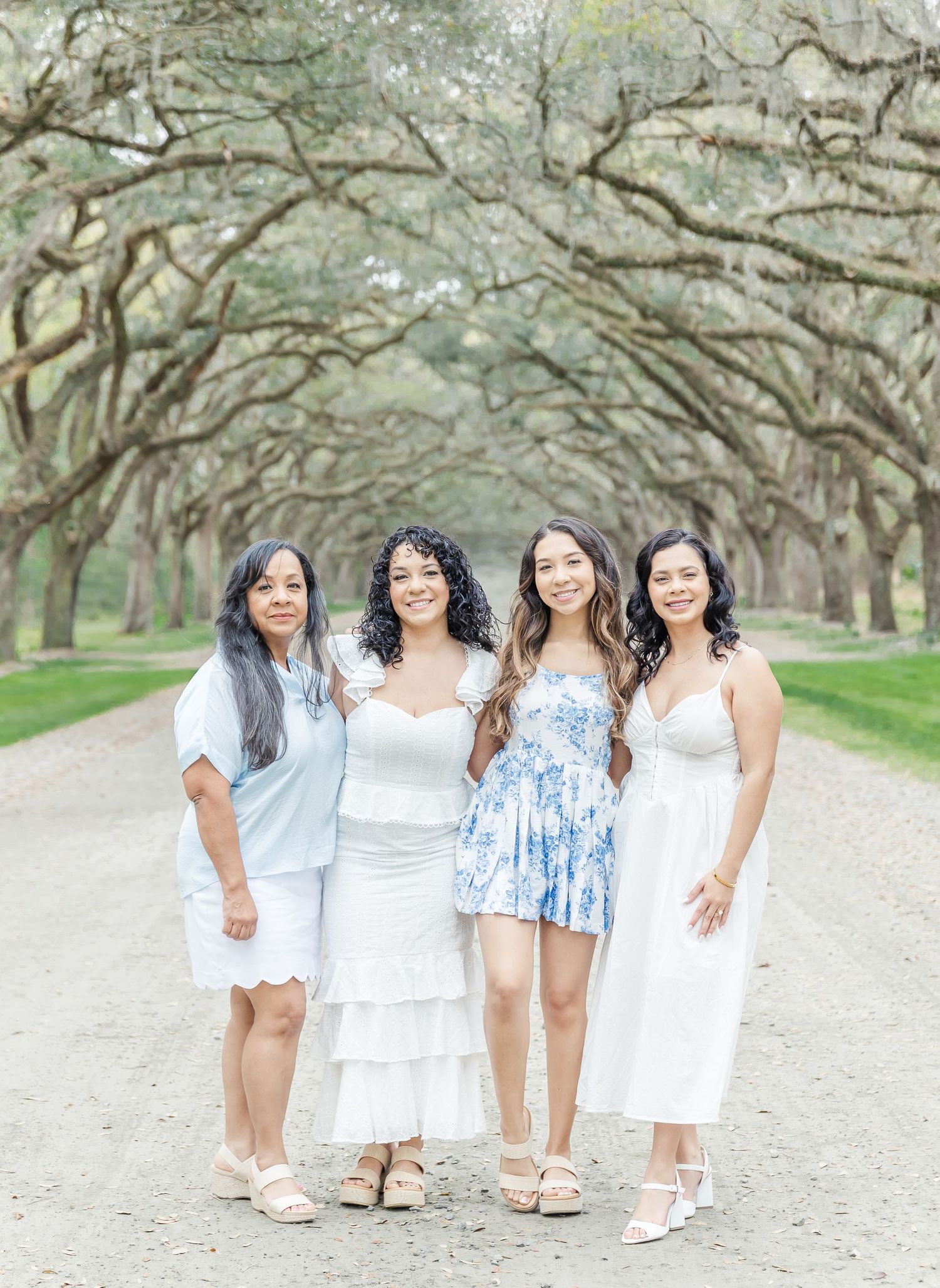 Four women in summer dresses stand together on a scenic path under arching oak trees.