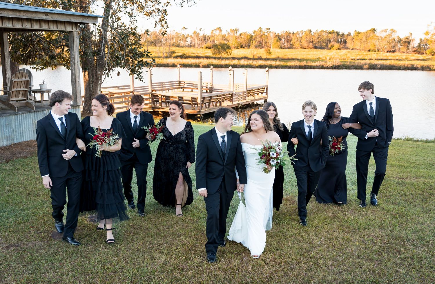 Wedding party walks together laughing across lawn with waterfront views.