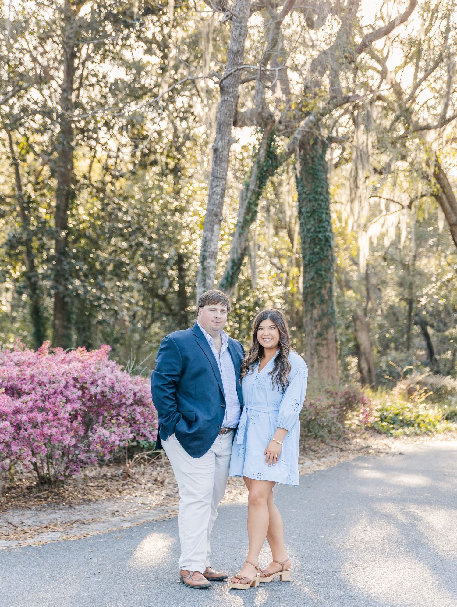 A couple poses on a sunlit path with blooming azaleas and trees in the background during a springtime photo session.
