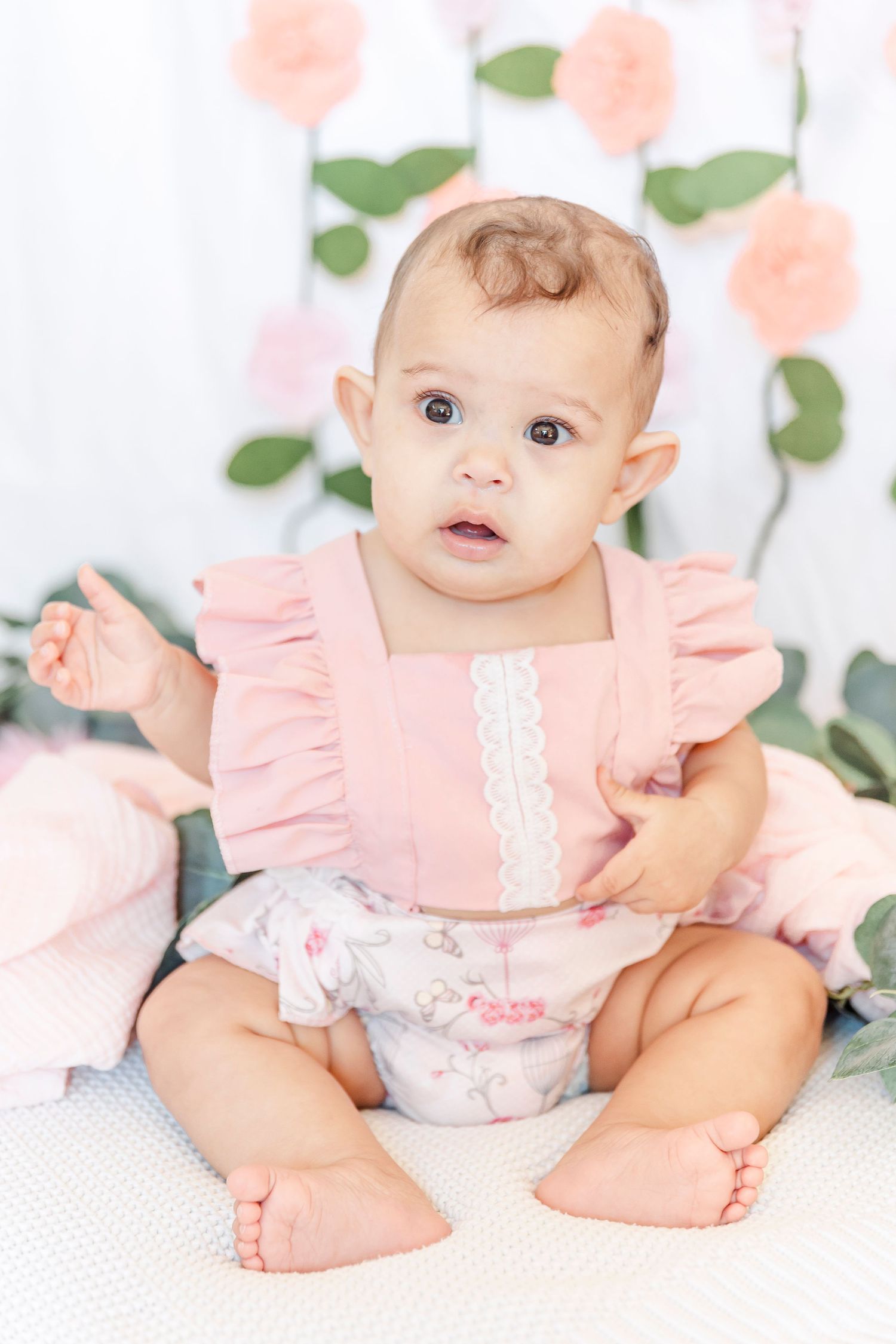 Baby in pink ruffled dress sitting against floral backdrop with soft peach roses.