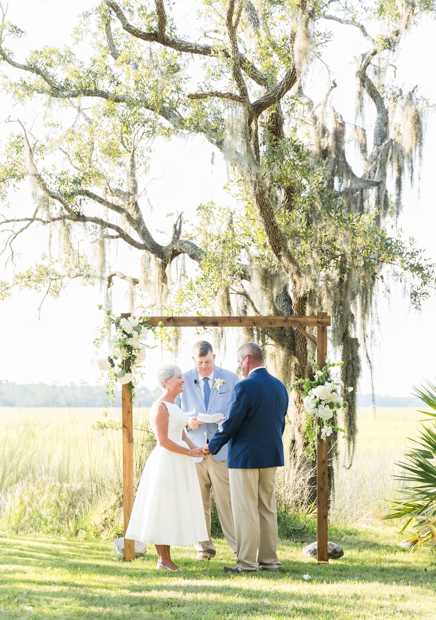 Wedding ceremony under rustic wooden arch with Spanish moss trees and marsh views.